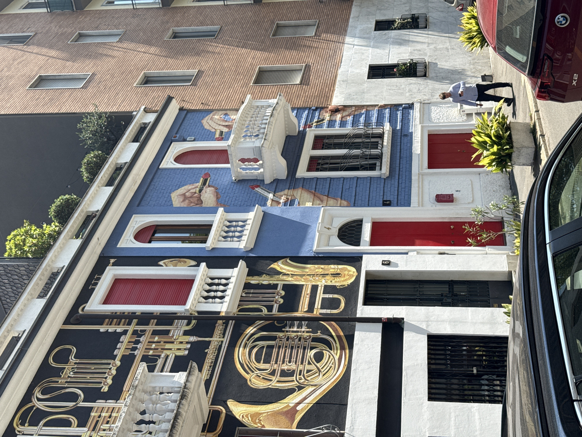 A mural facade graces a building in Milan, Italy, displaying trompe-l'oeil imagery of houses. The mural is divided into sections, each resembling a separate house with painted details like red doors, window bars, and white trim. The top section showcases lipstick tubes and hand details, while the bottom section features brass musical instruments like a French horn and trumpets. The mural is interrupted by architectural elements of the actual building, such as real windows and doorways. A man is walking on the sidewalk in front of the building, and two vehicles are parked nearby. The upper portion of the building exhibits traditional windows in a brick facade.