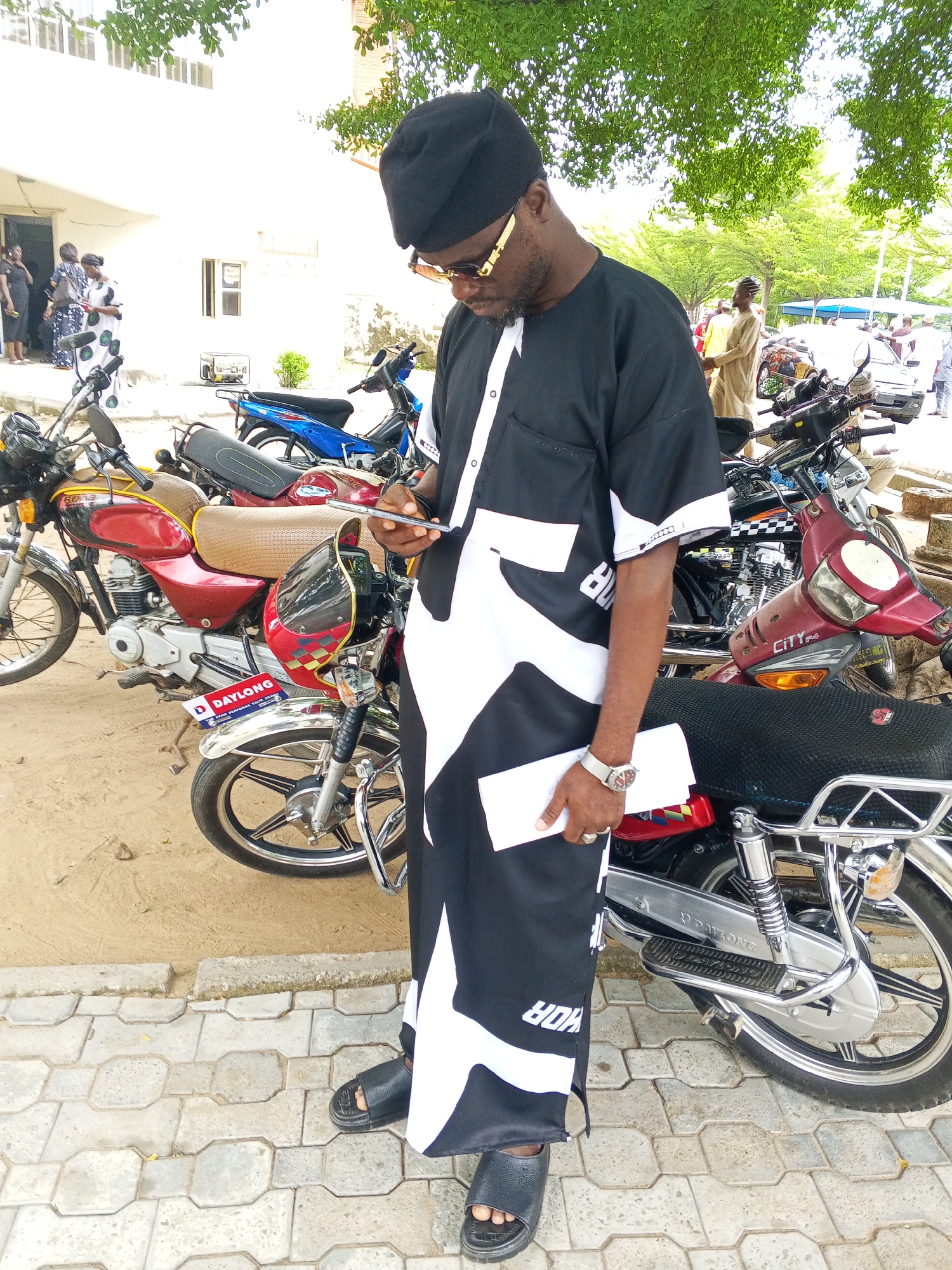 A man wearing a black cap, sunglasses, and a black and white patterned robe stands looking down at his phone, holding a white envelope in his other hand. He is in Lokoja, Nigeria, in an outdoor area with several motorcycles parked behind him. The motorcycles are of various colors, including red, blue, and black. In the background, there is a white building with windows and some trees. Other people are visible in the distance, some standing near the building and others near tents. The ground is a mixture of sand and patterned paving stones.