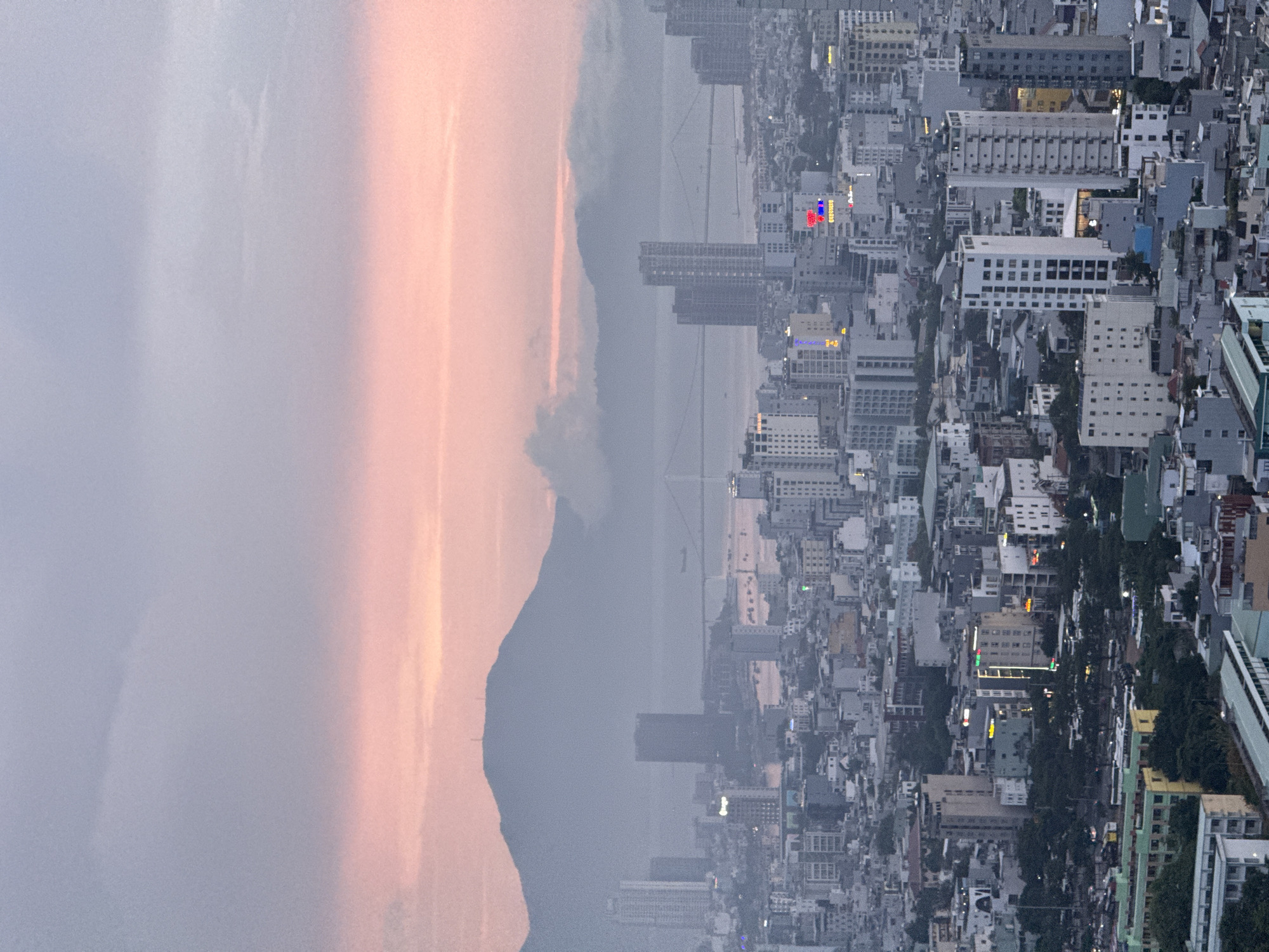 This is a high-angle aerial photograph of a city skyline at dusk. The sky is a soft blend of pale blue and pink, with the pink hues suggesting a sunset or sunrise. In the background, a large, hazy mountain range is silhouetted against the sky. A suspension bridge spans a body of water in the mid-ground, connecting parts of the city.
The foreground and mid-ground are dominated by a dense urban landscape filled with numerous buildings of varying heights and architectural styles. Many of the buildings are modern high-rises, constructed with concrete and glass, and some are illuminated with colored lights, indicating it is evening. The streets are visible in places, with cars and trees dotting the scene. The overall impression is of a bustling, developed city.
Given the presence of the Dragon Bridge (visible in the background, spanning the Han River) and the characteristic coastal mountain range often seen from Da Nang, this image is likely of the city of Da Nang, Vietnam. The specific vantage point appears to be from a tall building or elevated area overlooking the central district. There are no identifiable people or specific actions taking place, the scene is a static view of the urban environment.
