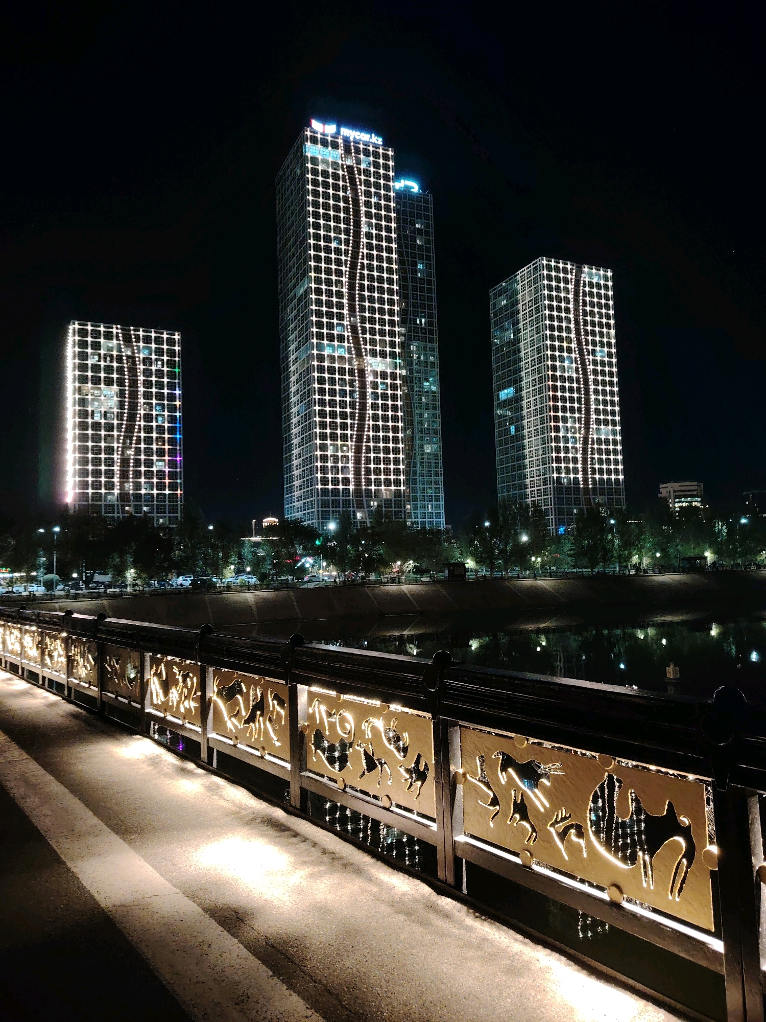 The image depicts a night scene in Astana, Kazakhstan, focusing on the "Northern Lights" residential complex, also known as "Khan Shatyr Residences," situated along the Ishim River. Three illuminated skyscrapers dominate the background, their facades decorated with intricate white light patterns resembling flowing waves. The tallest, central tower displays a logo on its peak that reads "mycar.kz" along with a national flag.
In the foreground, a pedestrian bridge with a decorative metal railing is prominent. The railing features golden cutouts of various animals, including camels, deer, and horses, backlit by warm white lights. The bridge's walkway is illuminated by strategically placed spotlights, casting long shadows. The Ishim River flows beside the bridge, reflecting the lights of the city. Trees line the riverbank, interspersed with streetlights and a few distant buildings. A street with cars is visible behind the trees, suggesting urban activity. The overall atmosphere is one of modern urban landscaping illuminated for nighttime viewing.