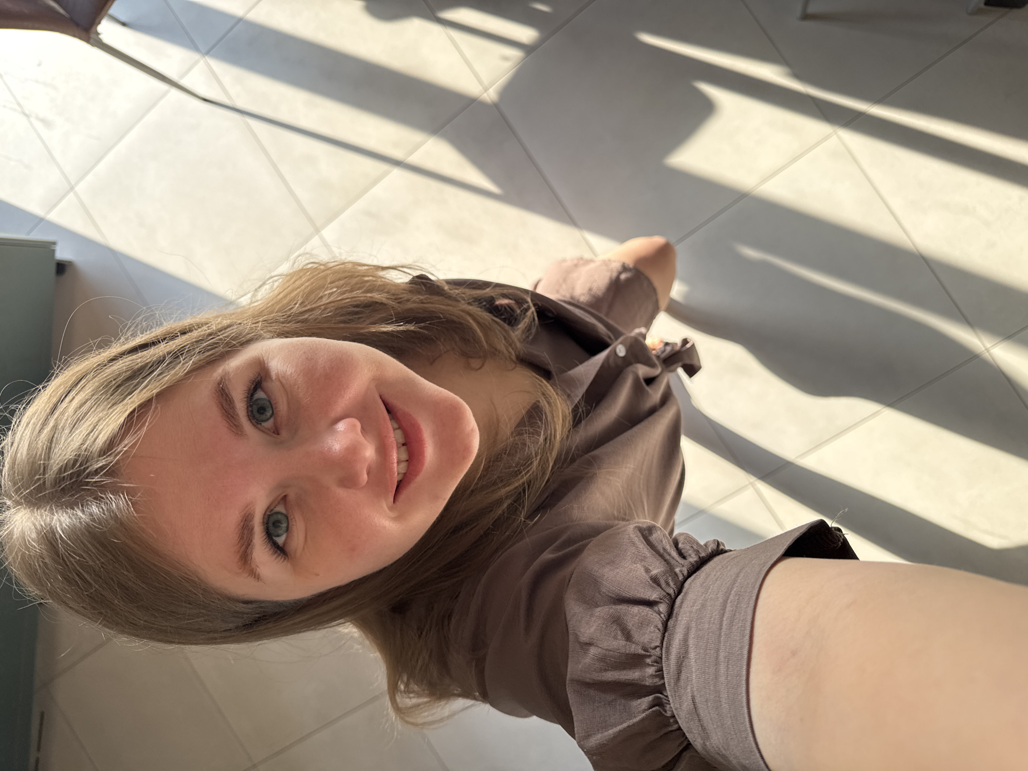 A selfie taken by a fair-skinned woman with blue eyes and light brown hair. She is smiling and wearing a brown long-sleeved blouse with gathered sleeves. The photo is taken from a high angle, looking down. The background consists of light-colored floor tiles with stark shadows cast by sunlight. The location appears to be indoors. The city specified is Ферма, Russia, but nothing in the image directly confirms this. The environment includes light-colored floor tiles and strong sunlight. There are no apparent objects or actions in the image beyond the woman taking a selfie in a brightly lit room.