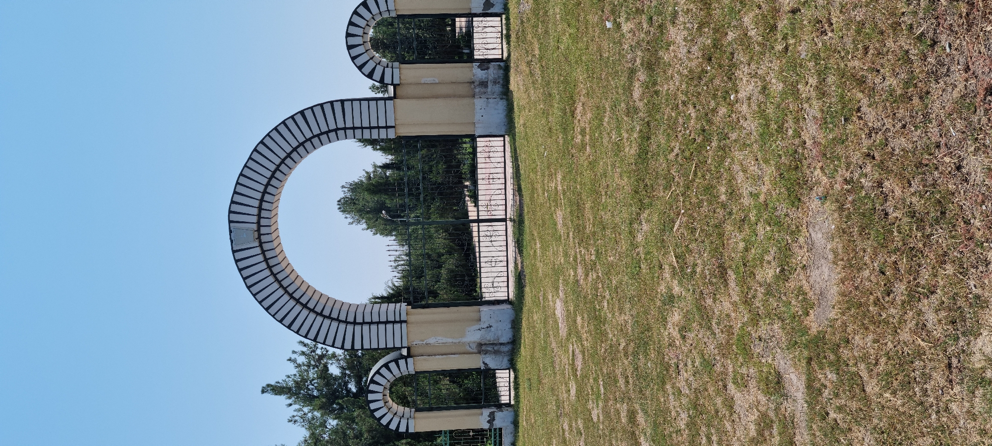 The image shows a park entrance with three arched gateways in the city of Ismoili Somoni, Tajikistan. The central archway is the largest, flanked by two smaller ones. The arches are decorated with black and white stripes, and the supporting pillars are yellow with weathered concrete bases. Behind the arches, a black metal fence encloses a wooded area with tall green trees. The ground in front of the entrance is covered with dry, sparse grass and dirt. The sky above is clear and blue. There are no people visible in the image.