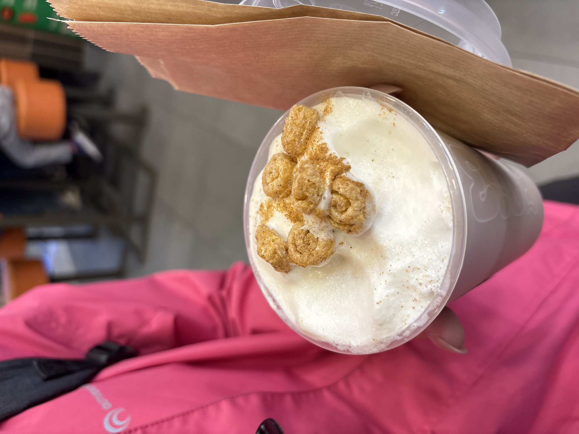 A high-angle, eye-level close-up shot shows a white drink topped with cereal, held against a pink jacket. The drink is in a clear plastic cup with a white lid, a brown paper sleeve around the cup, and topped with golden-brown circular cereal pieces and a light dusting of brown powder. The drink appears to be a latte or similar beverage with a frothy top. The background is blurry, but appears to be a store. The subject is wearing a bright pink jacket, and a portion of black bag is visible. The location is unspecified, but the mention of the city of Perm, Russia, indicates that the image may have been taken there.