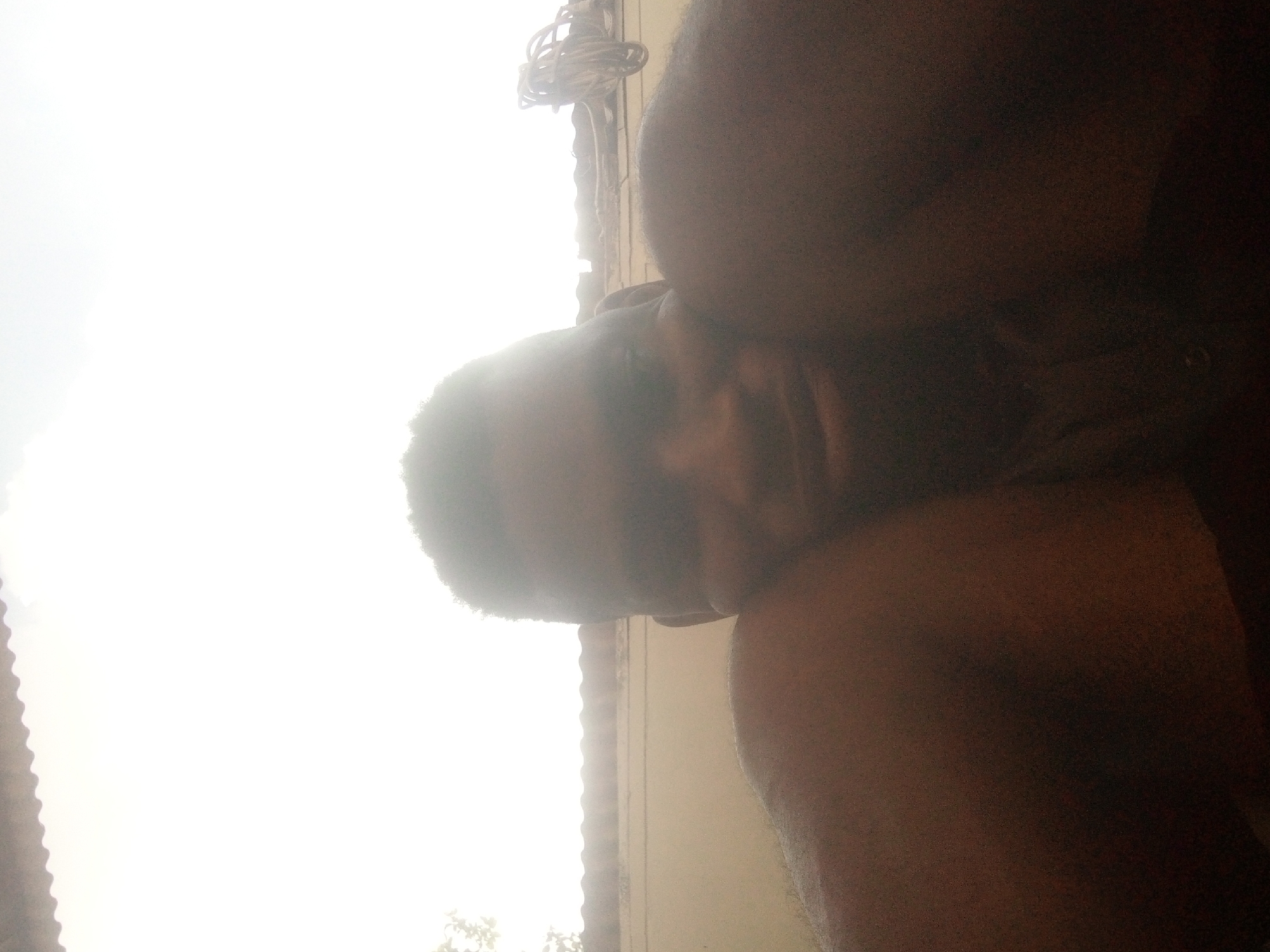 A close-up, tilted selfie of a Black male with short hair, illuminated by strong sunlight, obscures the facial details. A portion of his shoulder and chest are also visible. The backdrop appears to be a sun-drenched building in Ikorodu, Nigeria, inferred from the presence of architectural elements characteristic of the region. A small amount of electrical cables and leaves are visible.