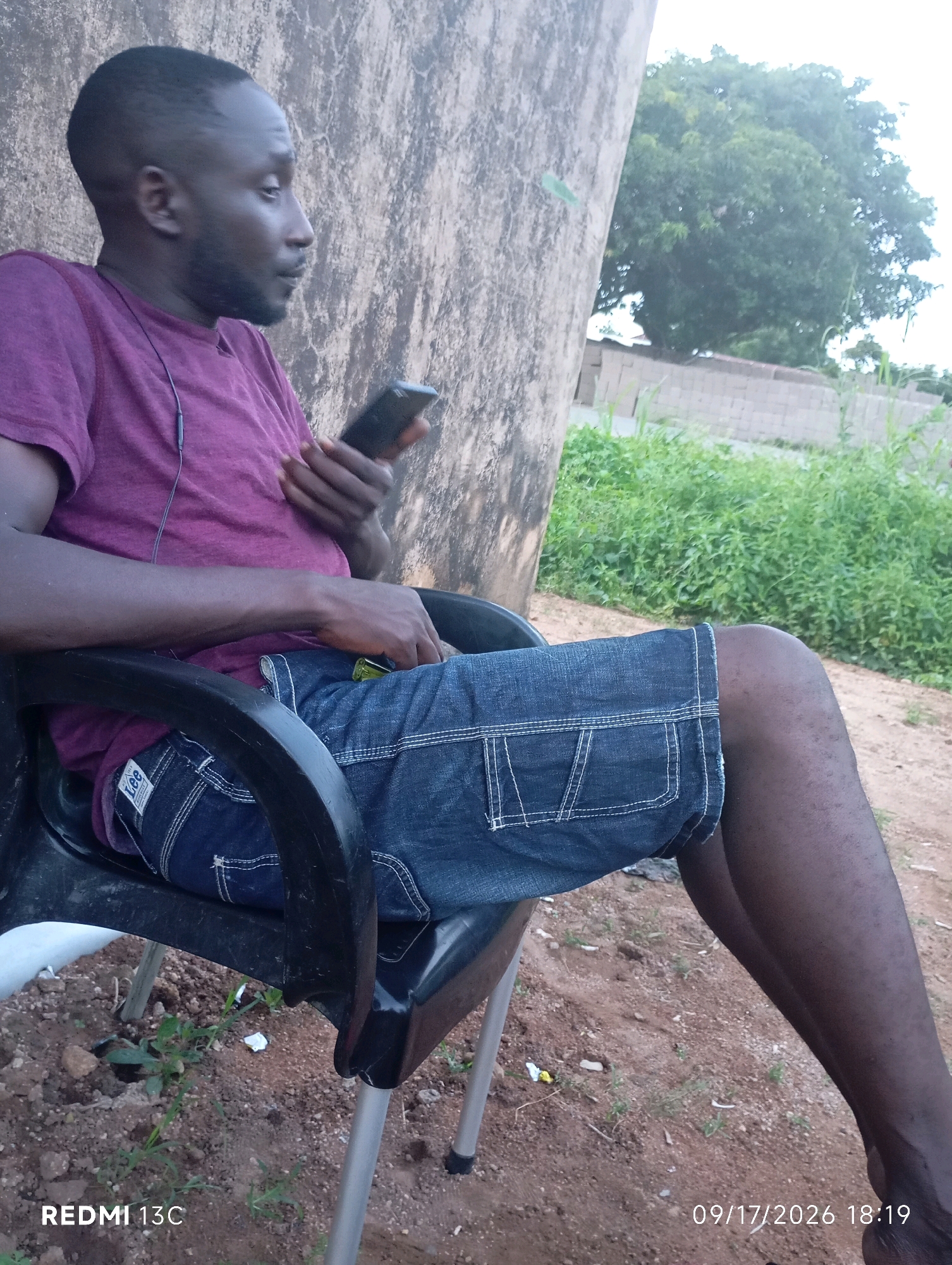 A man sits on a black plastic chair outdoors, his body angled to the right. He wears a purple t-shirt and denim shorts. In his right hand, he holds a black smartphone, and a thin black earphone cable runs from the phone to his ear. The man has dark skin and short hair, with a visible beard.
The background features a large, rough-textured tree trunk on the left, and dense green foliage on the right. The ground appears to be dirt and small rocks. The lighting suggests it is daytime, with soft, diffused light. A timestamp at the bottom right corner reads "09/17/2026 18:19". The camera branding "REDMI 13C" is visible at the bottom left. The scene is consistent with an outdoor setting in Shendam, Nigeria.