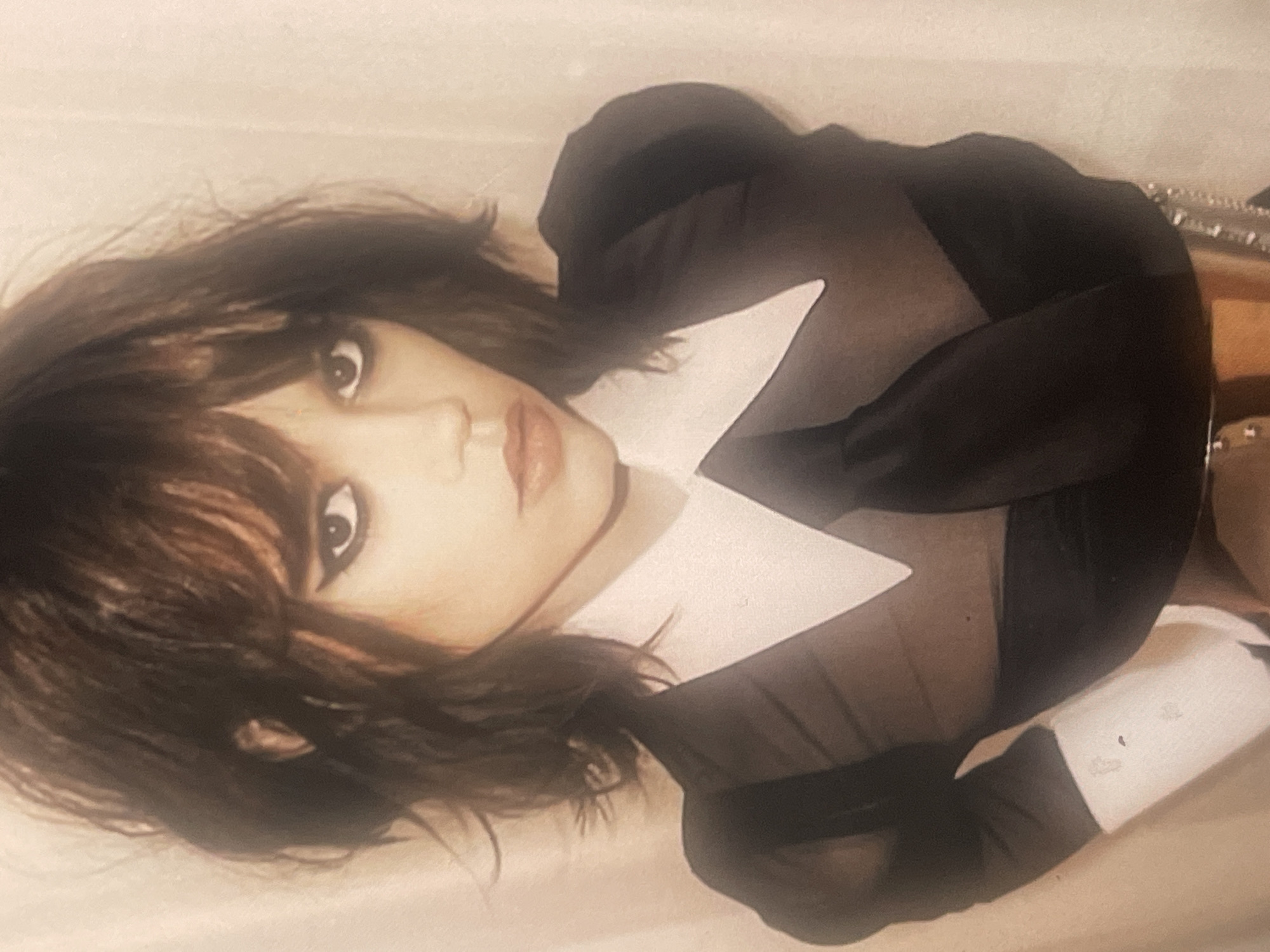 An indoor medium shot, rotated to appear vertical, captures a woman posing directly for the camera. She has dark, shoulder-length, layered hair with bangs that frame her face. Her eyes are accentuated with dark eyeliner and possibly eyeshadow, and her lips wear a light, glossy lipstick. She is wearing a dark, possibly sheer, top featuring prominent, sharply pointed white collar and white cuffs on puffy sleeves. A metallic chain or accessory is partially visible on the right side of the frame. The background is a plain, light, neutral-colored surface, which could be a wall or fabric. The woman's expression is direct and intense, with a serious gaze. The image has a soft, diffused lighting and a slightly grainy or textured quality. No text is visible in the image.
