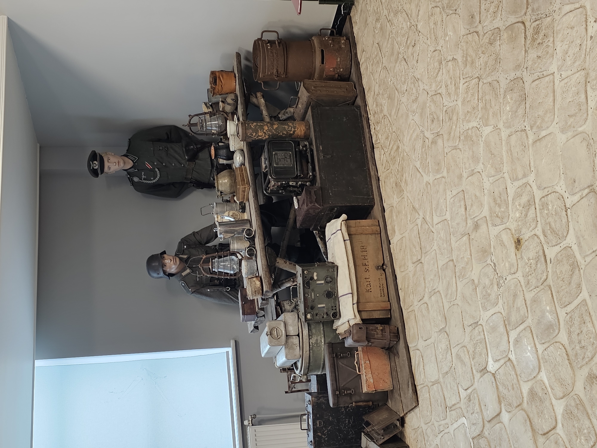 The image shows a museum display, likely in Архангельское, Russia, featuring two mannequins dressed in what appears to be World War II-era German military uniforms. One mannequin wears a helmet, while the other has a peaked military cap. They stand beside a shelf laden with various pieces of vintage military equipment. These include lanterns, metal canisters, wooden boxes, and communication devices such as radios. The backdrop is a plain gray wall on one side and a faux-stone wall on the other. A window is visible to the left.