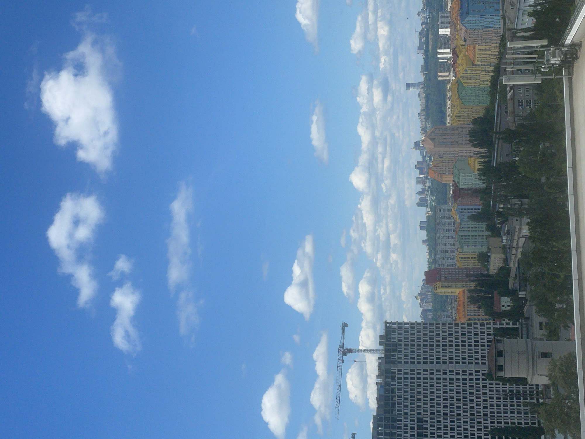 This is a medium-angle, eye-level outdoor shot of a city skyline under a bright blue sky with scattered white clouds. In the foreground, a grey, circular tower-like structure with a conical roof is visible, possibly a water tower or ventilation shaft. To its left, a massive, unfinished grey building with a grid-like facade dominates the scene, with a construction crane extending from its upper portion. Beyond this, a cluster of colorful residential buildings, painted in various hues of blue, green, yellow, orange, and red, stretches across the middle ground, forming a distinctive architectural landscape. These buildings appear to be mid-rise apartments. Further in the background, a hazy city skyline with numerous buildings of varying heights is discernible, and in the far distance, a tall, slender tower with a rounded top stands out against the sky. The overall scene depicts a developing urban environment in Kyiv, Ukraine, likely within the Obolonskyi district, known for its colorful residential complexes. No people are visible in the image.