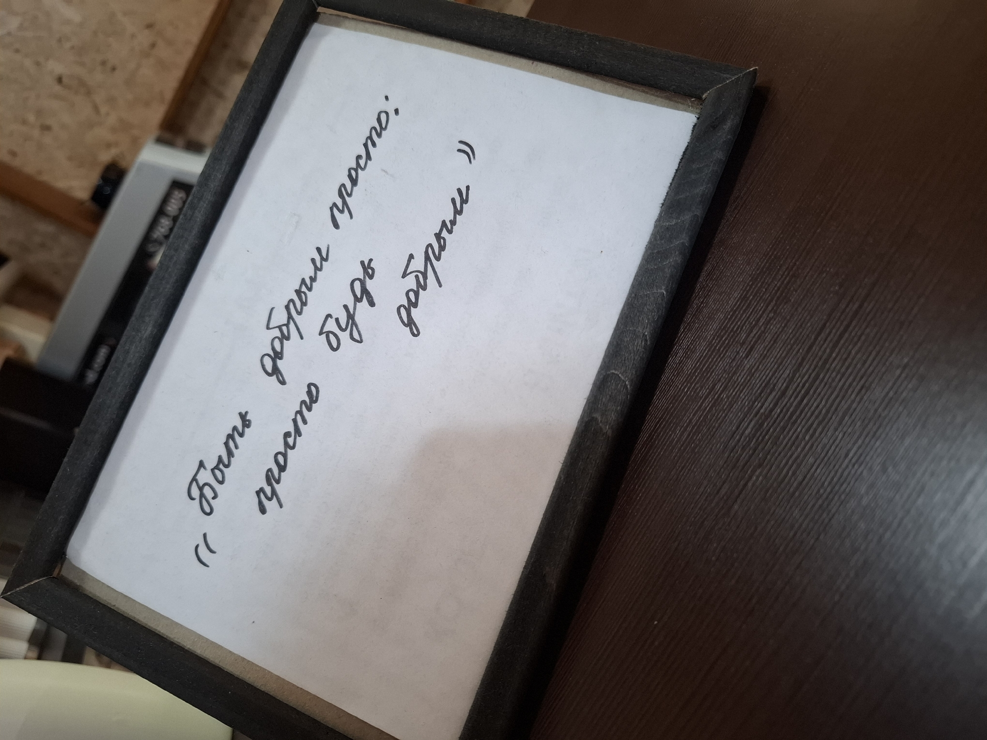 A framed handwritten quote is placed on a dark brown wooden surface. The quote, written in black cursive, reads "Быть добрым просто: просто будь добрым". The frame is dark gray and made of wood, with a visible grain pattern. The background shows a blurred out room with wooden paneling and some equipment. The overall scene suggests an indoor setting, possibly a cafe or office space. No people are present.