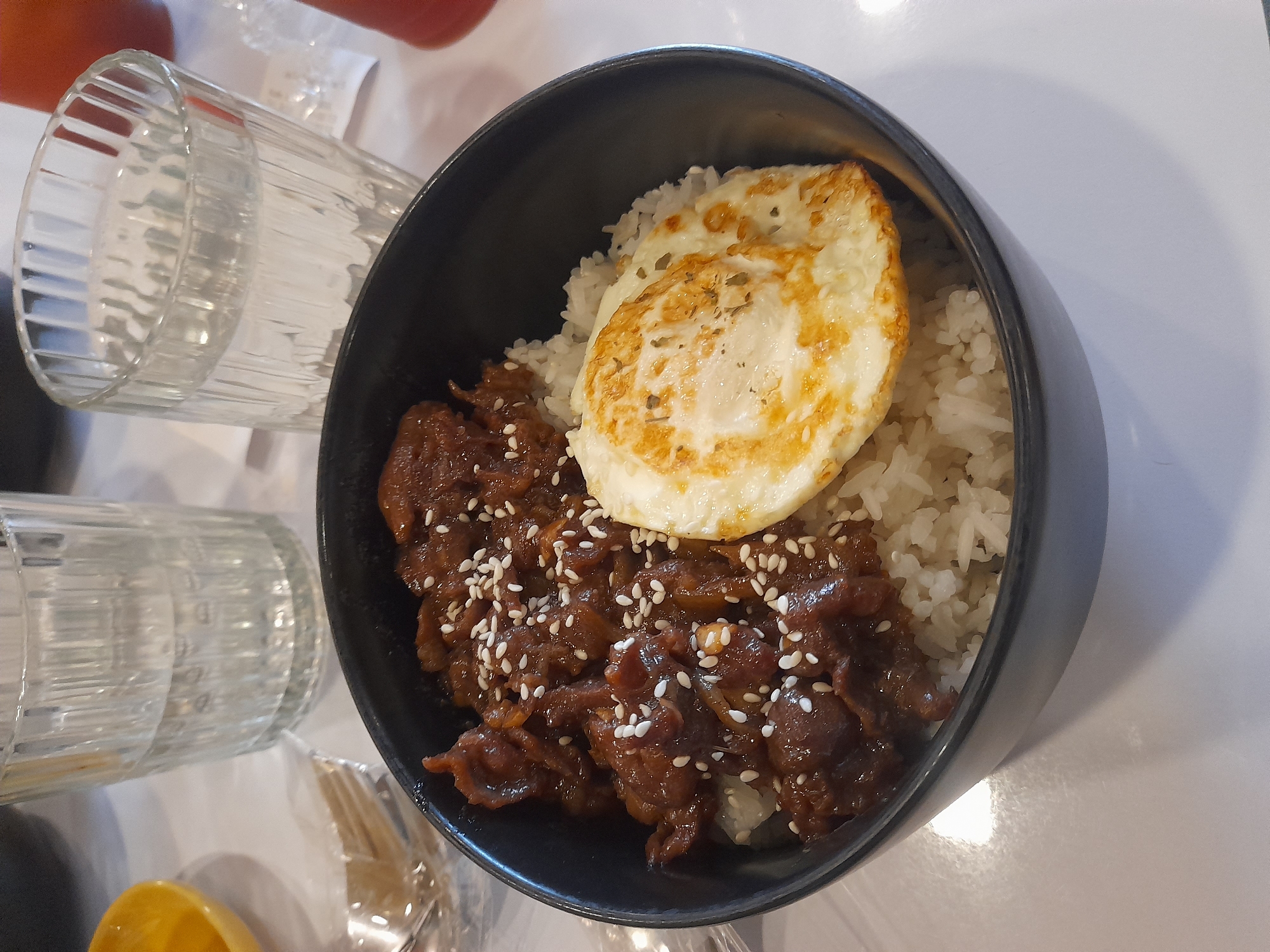 The image shows a bowl of beef teriyaki with rice and a fried egg on top, sprinkled with sesame seeds. Two glasses are partially visible on the left side of the frame. The setting is likely a restaurant in Surabaya, Indonesia. The bowl is on a white table.