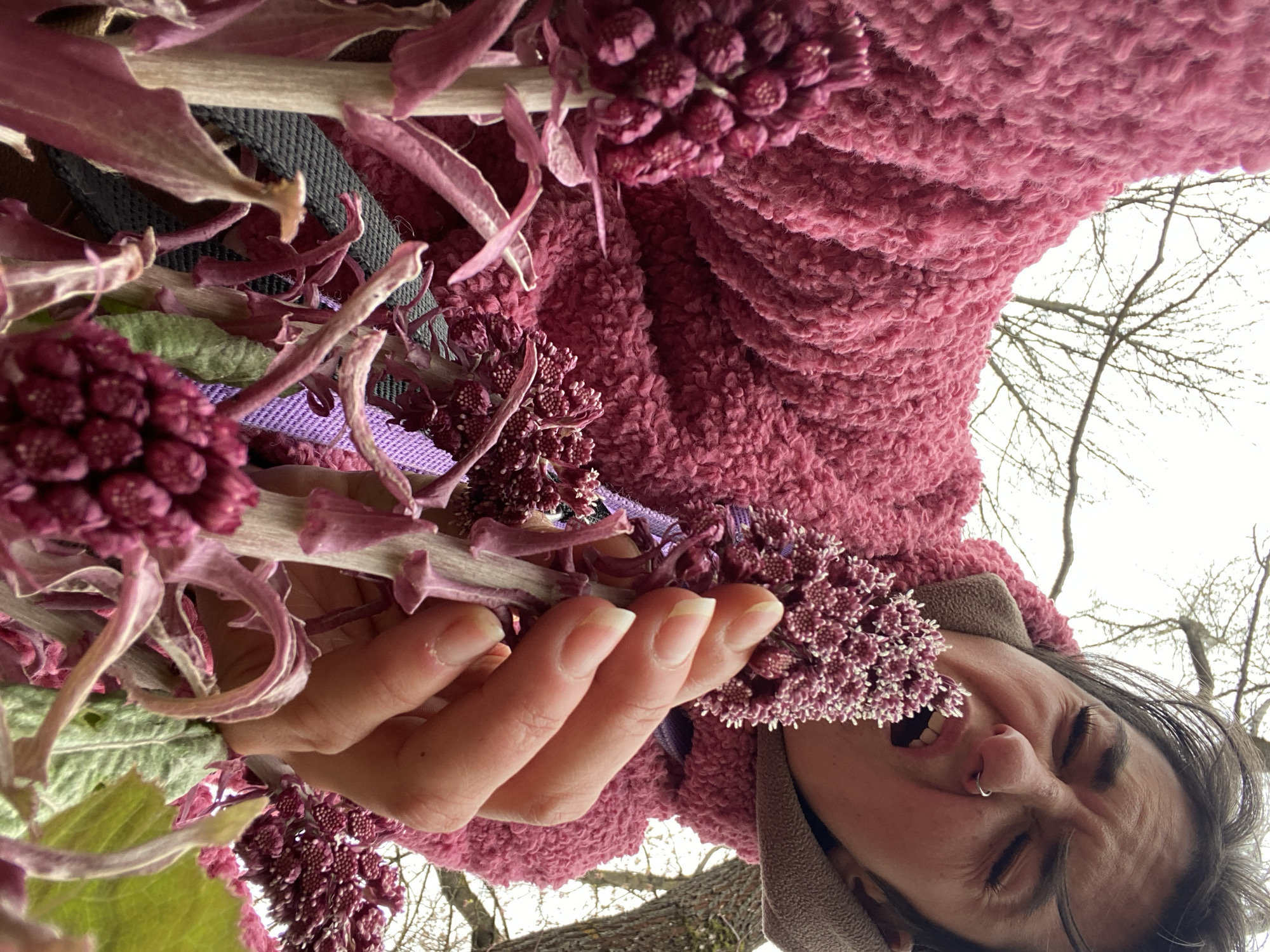 A person is seen from a low angle holding a bunch of purple flowers in their left hand. The person is wearing a pink, fluffy jacket and has a nose ring. They are smiling with their eyes closed. A tree with bare branches is visible in the background. There is no clear indication of Moscow, Russia, in this image.