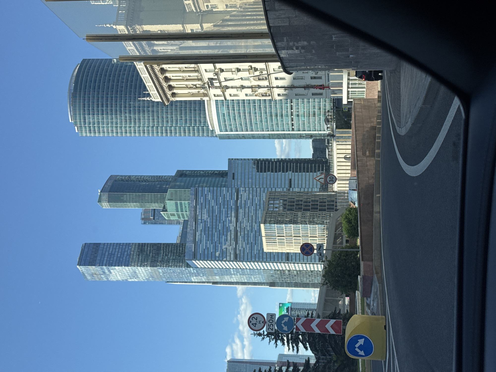This is a vertical, medium-shot photograph taken from inside a vehicle looking out at a city street scene in Moscow, Russia. The image is dominated by a cluster of modern skyscrapers, their glass facades reflecting the bright blue sky and scattered white clouds. The tallest buildings are a series of interconnected towers with angular, geometric designs. In the foreground, a dark asphalt road curves to the right, bordered by a white lane marking. Several road signs are visible, including a circular sign with a red border and the number "30" inside, indicating a speed limit, and a sign with two opposing arrows, suggesting a lane change or traffic direction. A yellow traffic bollard is also present.
To the left of the skyscrapers, an older, ornate building with classical architectural elements stands out. Some of the skyscrapers appear to have a green tint from their glass exteriors. The reflection in the glass of the older building reveals more of its detailed facade. The time of day appears to be midday or early afternoon, given the bright sunlight and clear skies, suggesting fair weather. The overall impression is one of a bustling urban environment, showcasing a mix of contemporary and older architectural styles. There is no visible activity of people.