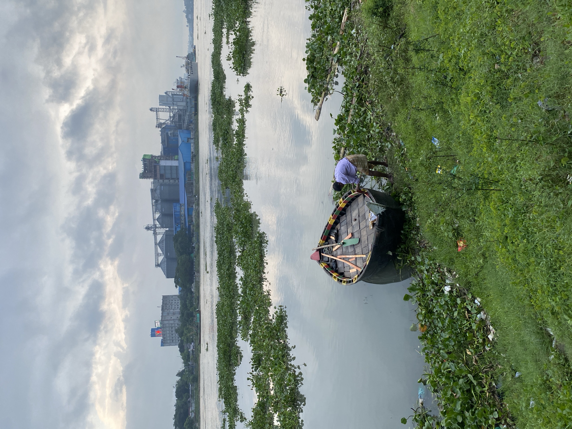 The image depicts a view of the Buriganga River in Dhaka, Bangladesh, with an industrial complex visible in the background. In the foreground, a man is standing next to a small boat, appearing to be tending to it. The boat is partially submerged and filled with oars. The river is covered in dense patches of water hyacinths, with some visible litter along the embankment. The industrial complex in the distance features several large silos and buildings, suggesting a port or manufacturing facility. The sky is overcast, contributing to a diffused, muted lighting of the scene.