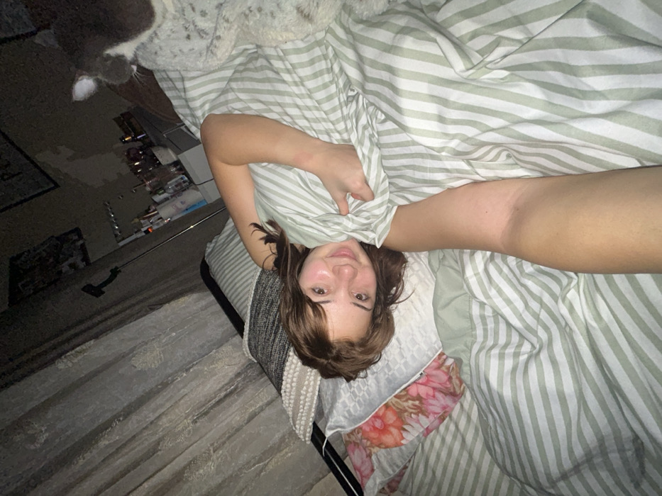 A girl is lying in bed with green and white striped sheets pulled up around her. She faces the camera with a slight smile. Her hair is brown with bangs. Behind her, a floral patterned pillow sits on the bed. In the background, there's a dimly lit room with a rug, a table with items on it, and a cat partially visible in the top left corner. The image appears to have been taken at an angle, giving a slightly distorted perspective.