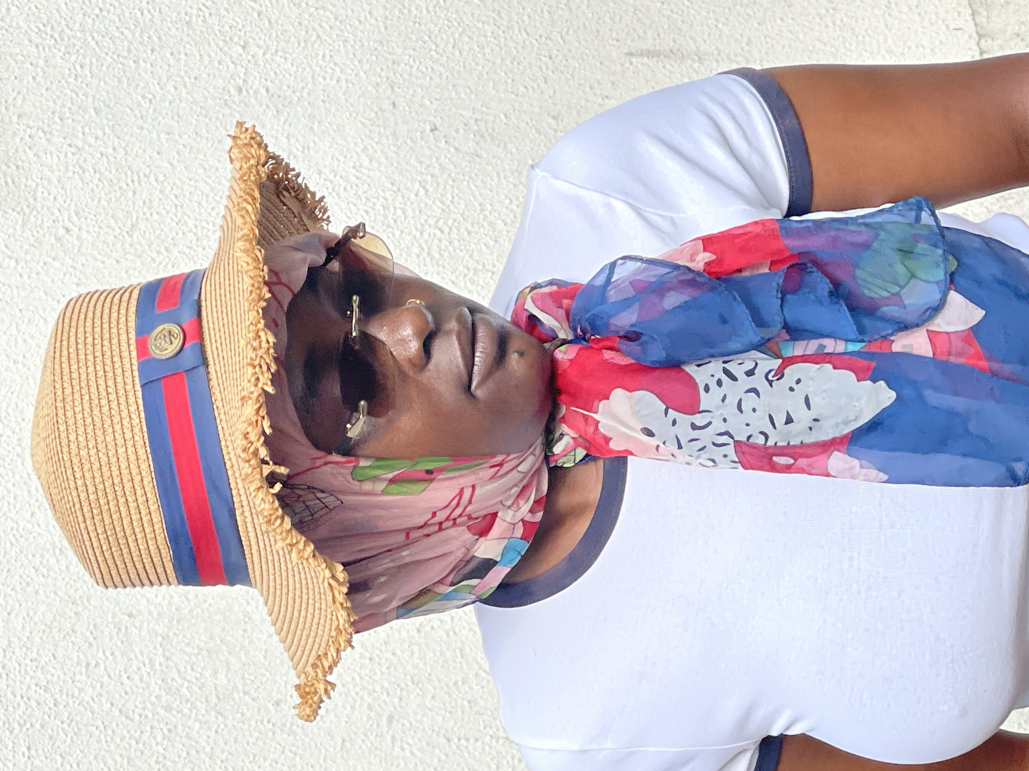 A medium shot depicts a dark-skinned individual, angled slightly, wearing a straw hat with a blue and red striped band, sunglasses, and a multi-colored scarf tied around their head and neck. They are wearing a white shirt with dark trim around the neck and sleeves. The background appears to be a textured white wall. While the image context suggests the location is Gwarinpa, Nigeria, there are no visual elements to confirm this.