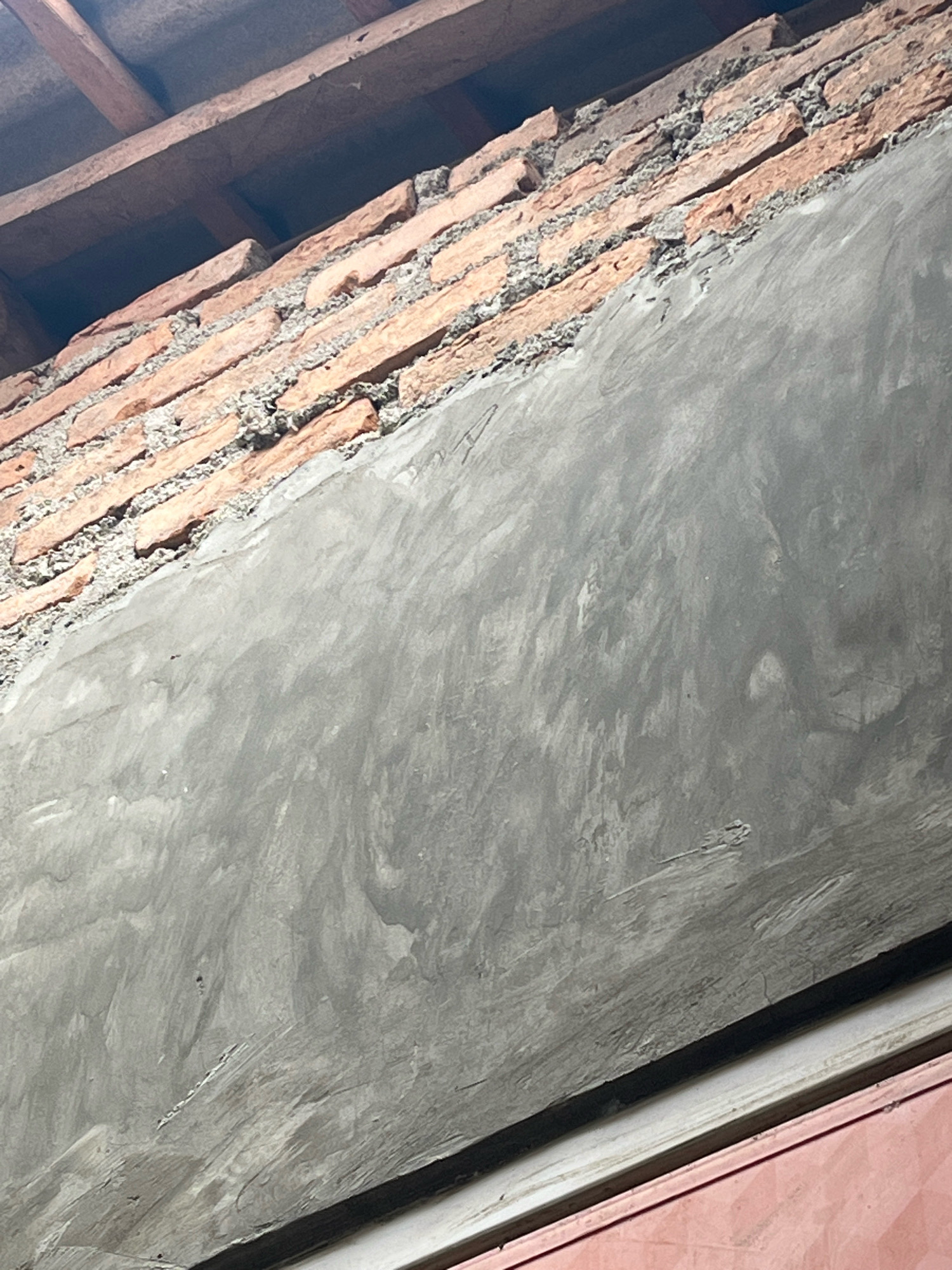 The image shows a close-up of a brick wall under construction, with a section already plastered. The top of the image reveals wooden beams that appear to be part of a roof structure. The bricks are reddish-orange and laid with visible mortar. The plaster is gray and has a rough, textured finish. The image is likely from Kebumen, Indonesia, as indicated by the context provided. There are no people or specific actions depicted.