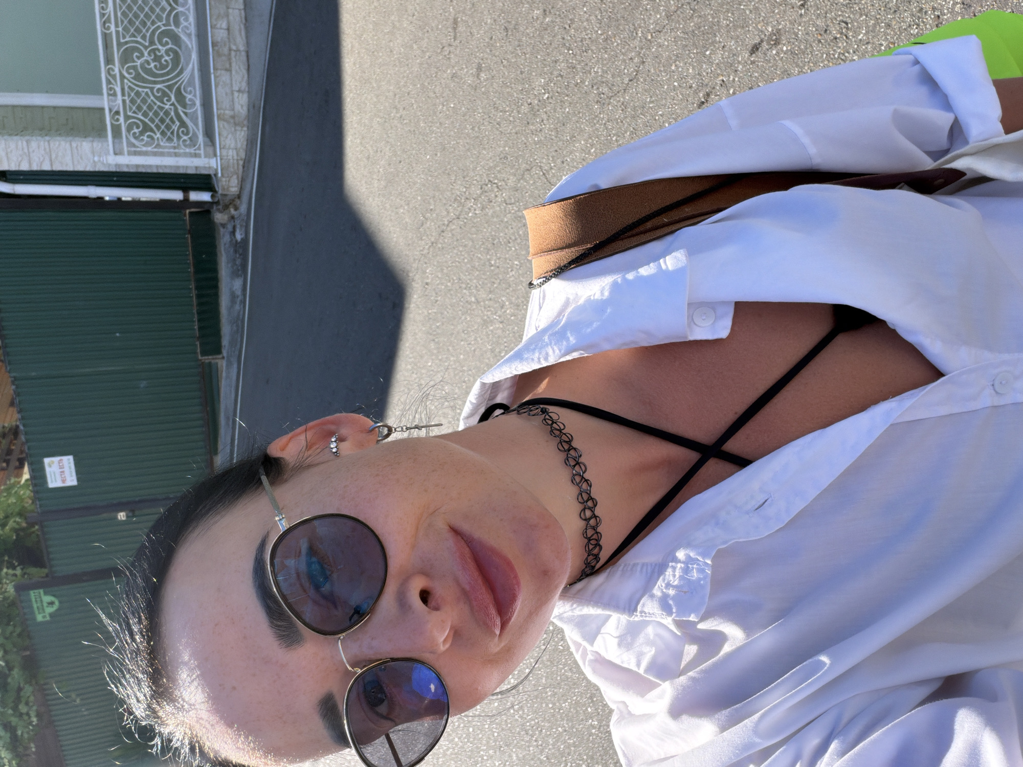 A woman is taking a selfie outdoors, with a slightly blurry street and buildings in the background. She has dark hair, is wearing round sunglasses, a white open button-up shirt over a black top, a black choker necklace, and a crossbody bag with a tan strap. Her left ear has a silver stud earring and a dangling cross earring. The scene appears to be in Sochi, Russia, based on the surrounding architecture and vegetation.