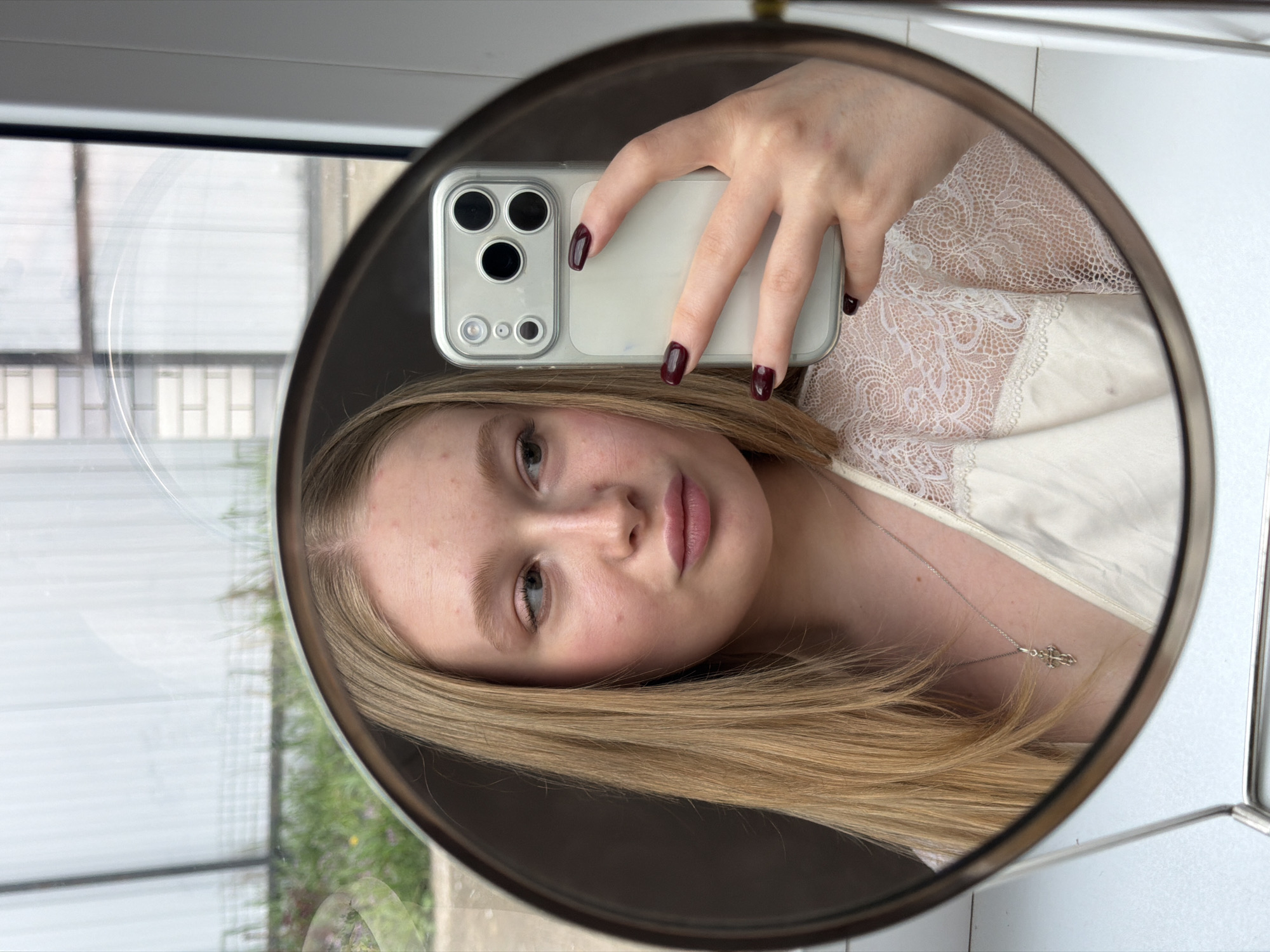 A young, fair-skinned woman with light blonde hair is taking a selfie in a round mirror. She is holding a smartphone in her right hand, its camera lens and flash visible. She is wearing a white lace-trimmed top and a delicate silver necklace. The reflection in the mirror shows a window with a view of greenery outside, suggesting an indoor setting. The photograph could have been taken in Rossiyskiy, Russia, but there is no concrete information to confirm this location definitively.