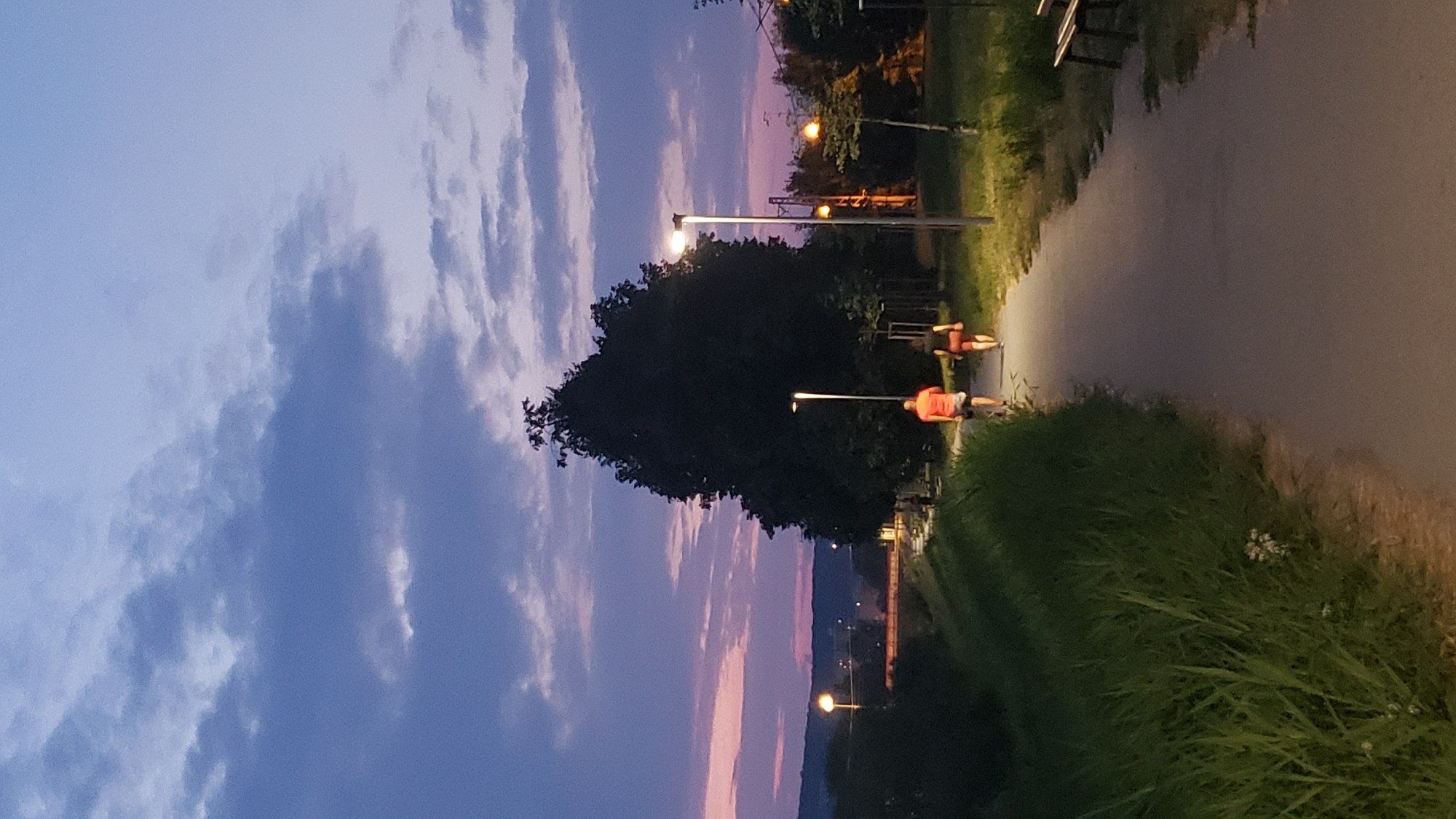 The image depicts a paved pathway in a park at dusk in Prešov, Slovakia. Two individuals are walking away from the camera on the path. The person in front is wearing an orange t-shirt and shorts, and the person behind them is wearing a dark t-shirt and shorts. They are silhouetted against the lit path and surrounding vegetation. Several lampposts line the pathway, some illuminated, casting light on the scene. A large, dark tree stands to the right of the pathway, and a dense patch of tall green grass is visible to the left. In the background, a softly lit landscape with rolling hills is visible under a twilight sky with scattered clouds tinged with pink and purple hues. Distant city lights are also visible on the horizon.
