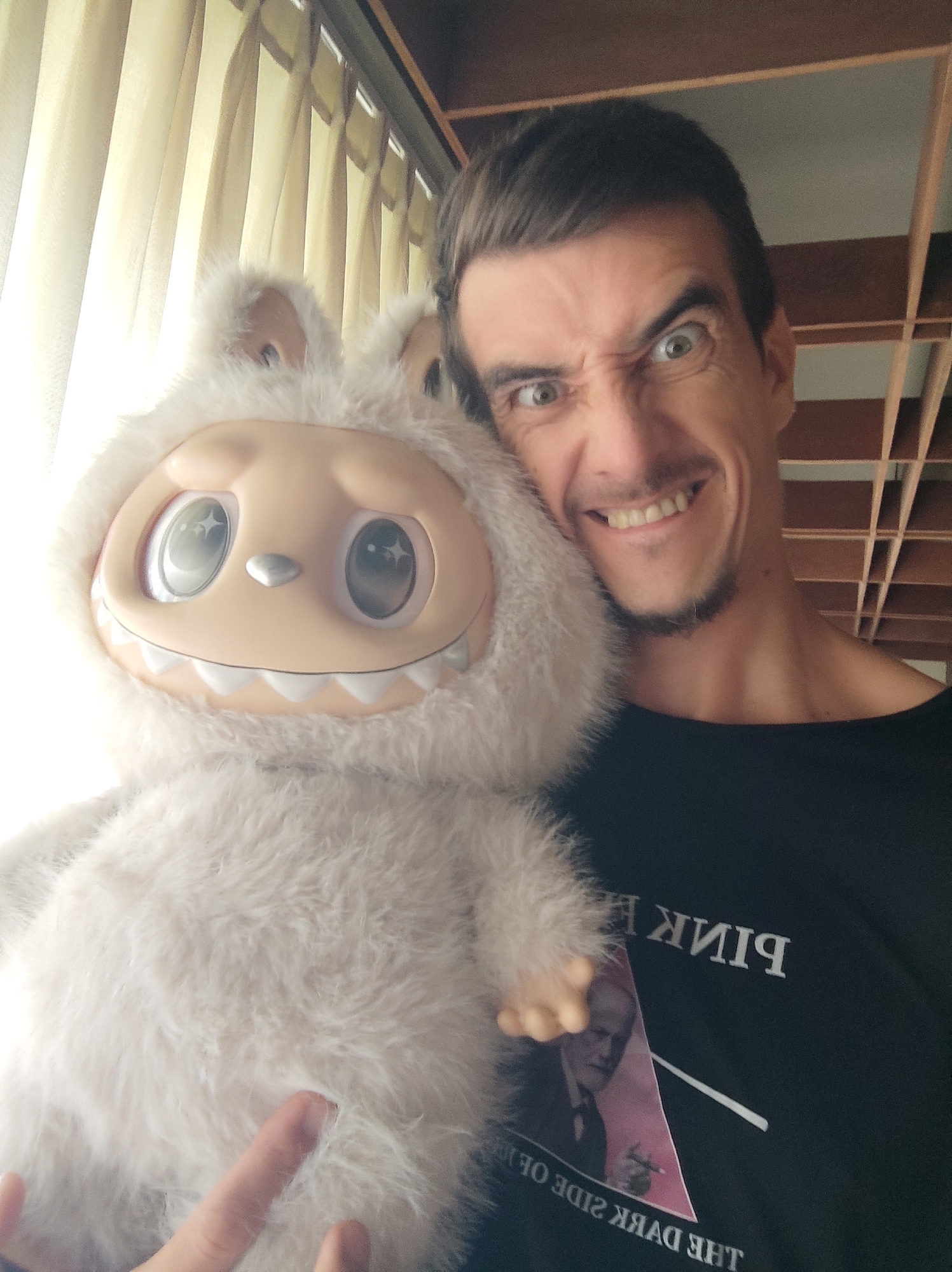 An indoor selfie captures a man embracing a large, fluffy, light-colored plush toy. The man, with short dark hair and a goatee, is making a humorous, exaggerated snarling face at the camera, with wide green eyes and a visible grin. His left hand is seen resting on the plush toy.
The plush toy, resembling a yeti or monster, has large, sparkling eyes with star-shaped reflections and a wide, fixed grin displaying a row of white teeth. Its body is covered in soft, white or light gray fur.
The man is wearing a black t-shirt with partially visible, horizontally reflected text. The word "PINK" is visible at the top, followed by an image of a man (possibly Sigmund Freud) within a pink triangle. Below this, the reflected phrase "THE DARK SIDE OF THE" can be read, strongly suggesting a reference to Pink Floyd's "The Dark Side of the Moon" album.
The background on the left is brightly lit by natural light coming through light-colored curtains, indicating daytime. To the upper right, a wooden slatted structure, possibly part of a ceiling or roof, is visible, consistent with architecture found in locations like North Kuta, Indonesia. The overall scene conveys a playful and jovial atmosphere.