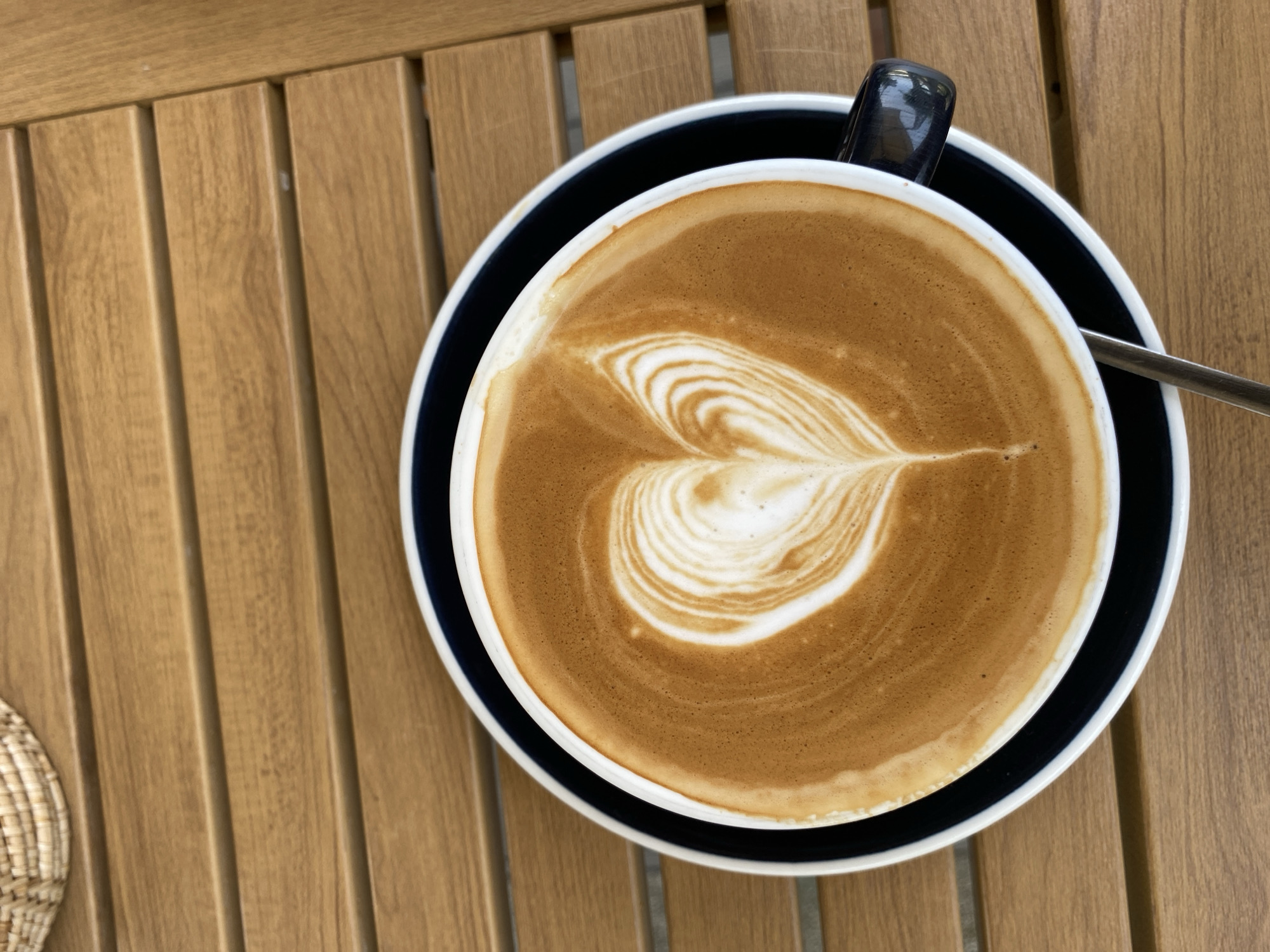 A top-down shot displays a cup of latte art with a heart design on a wooden slatted table. The coffee sits in a white cup on a dark blue saucer with a thin white stripe at the saucer's edge. A spoon is partially visible on the right side of the cup. A small piece of woven basket is on the bottom left corner. The image was likely taken outdoors at a cafe or restaurant in Choeng Thale, Thailand. No people are visible.