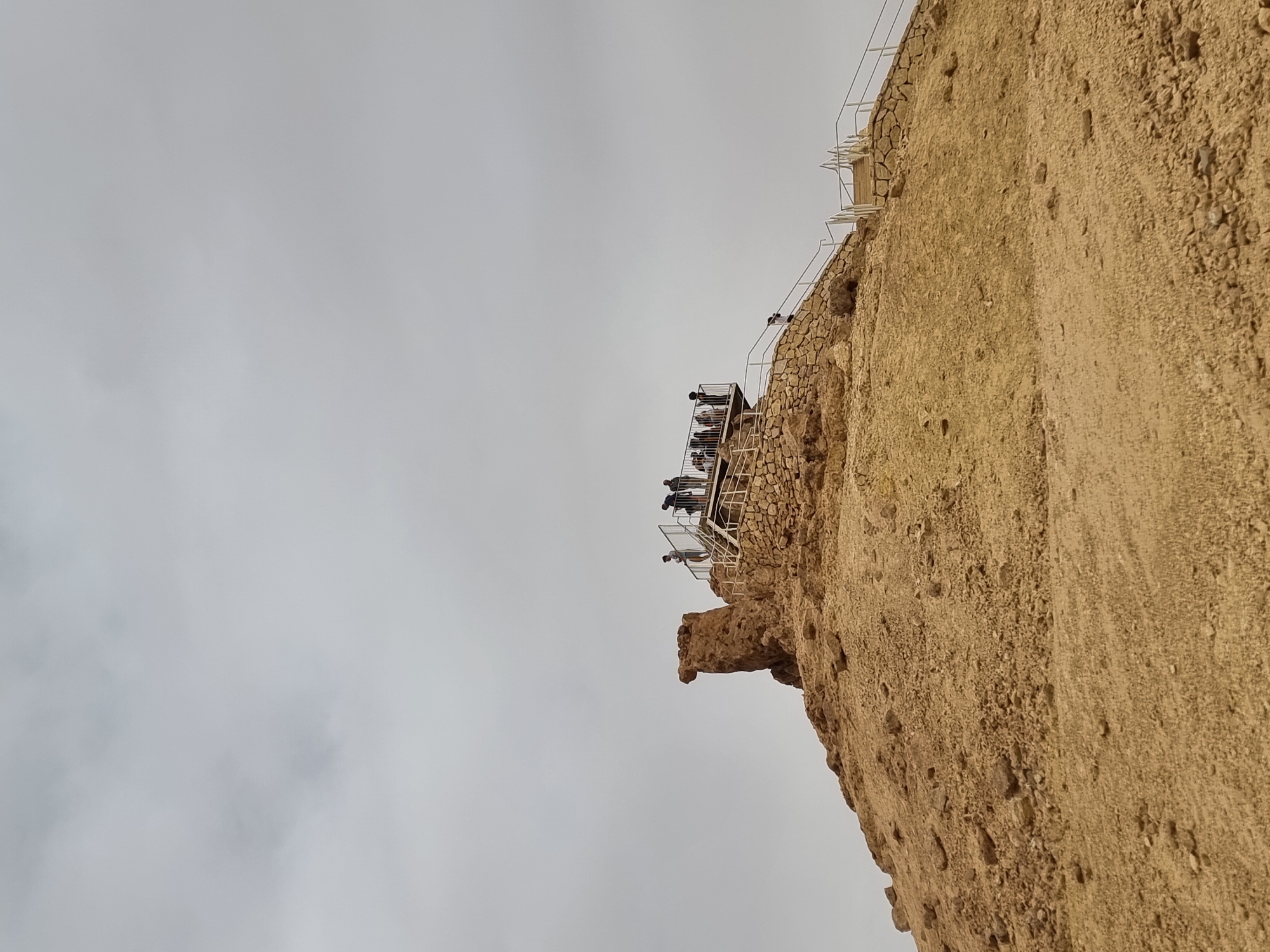 The image depicts a cliffside structure located in Mitzpe Ramon, Israel. Atop the cliff, a viewing platform with a white metal railing is populated by several people, estimated to be around a dozen. The cliff face is composed of layered rock and soil, showing a mix of textures. The sky is overcast and grey. The image is taken from a low angle, emphasizing the height and verticality of the cliff.