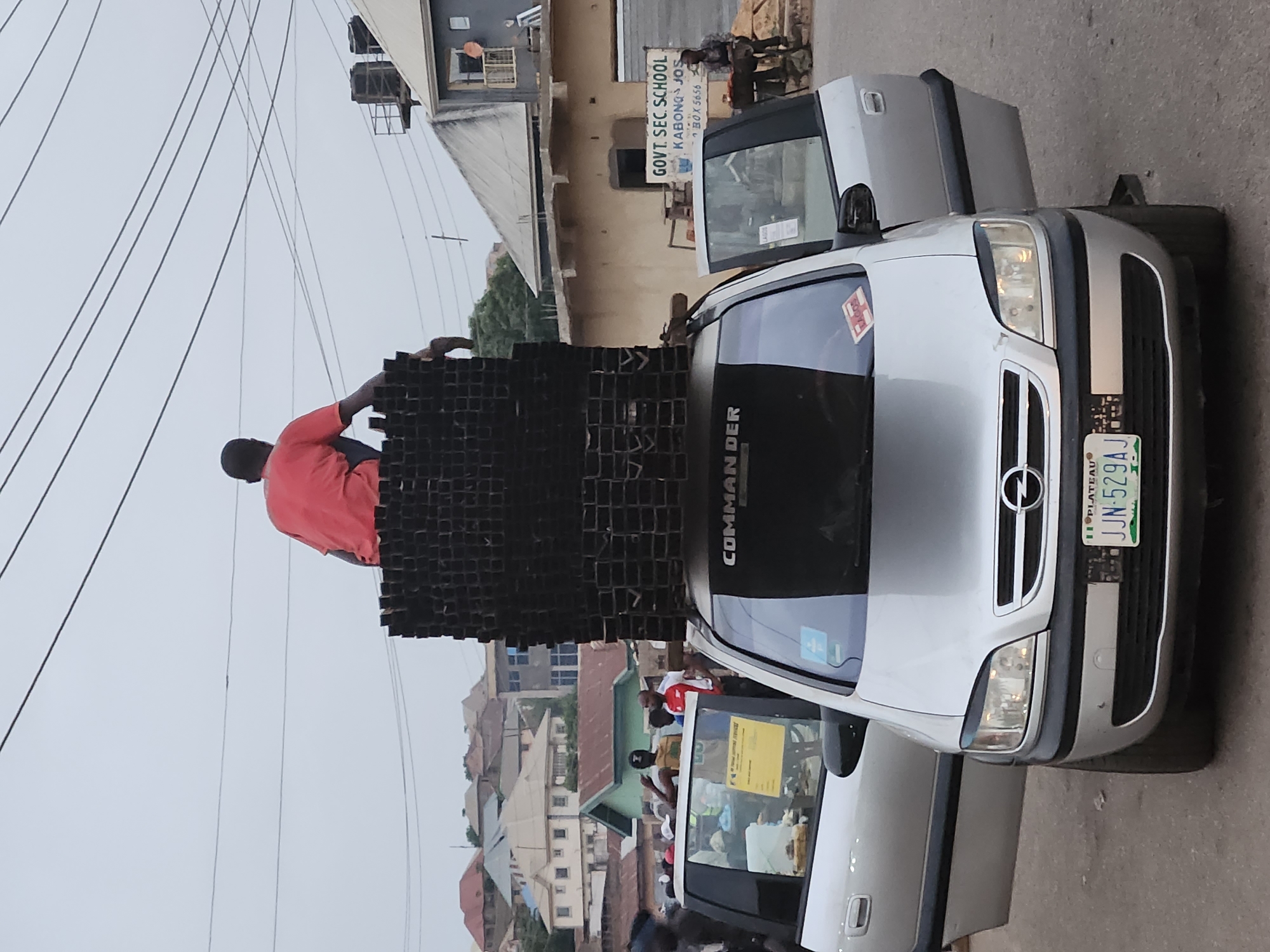 A man in a red shirt is standing on top of a large stack of rectangular metal tubes on the roof of a silver Opel Zafira. The car has a license plate that reads "JJN-529AJ 11 PLATEAU LAGOS". The rear window of the car has the word "COMMANDER" written across it. The car is parked in front of a building with a sign that reads "GOVT. SEC. SCHOOL KABONG JOS". Other buildings are in the background, indicating that the photo was taken in a built-up area in Kabong, Nigeria. Other people are standing near the car. Overhead power lines are also present.