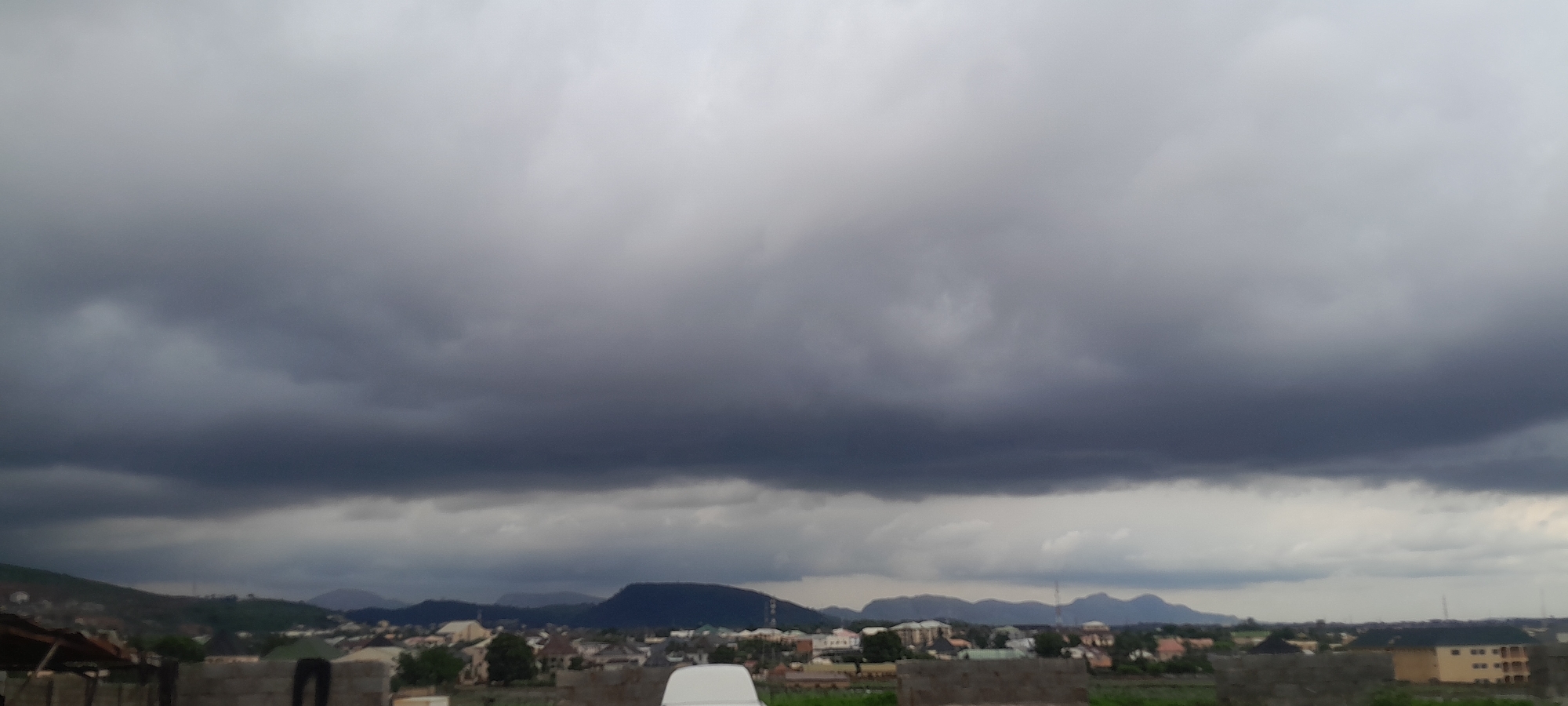 The image depicts a city scene in Abuja, Nigeria, under a heavy, dark cloud cover. The sky is dominated by ominous, grey clouds, suggesting an impending storm. Below the clouds, a range of hills and mountains can be seen in the distance. In the mid-ground, a settlement with various buildings, including houses and larger structures, is visible. The foreground features a partially constructed brick wall and a white object, possibly a vehicle or furniture, which is out of focus. The overall atmosphere is somber due to the dark sky.