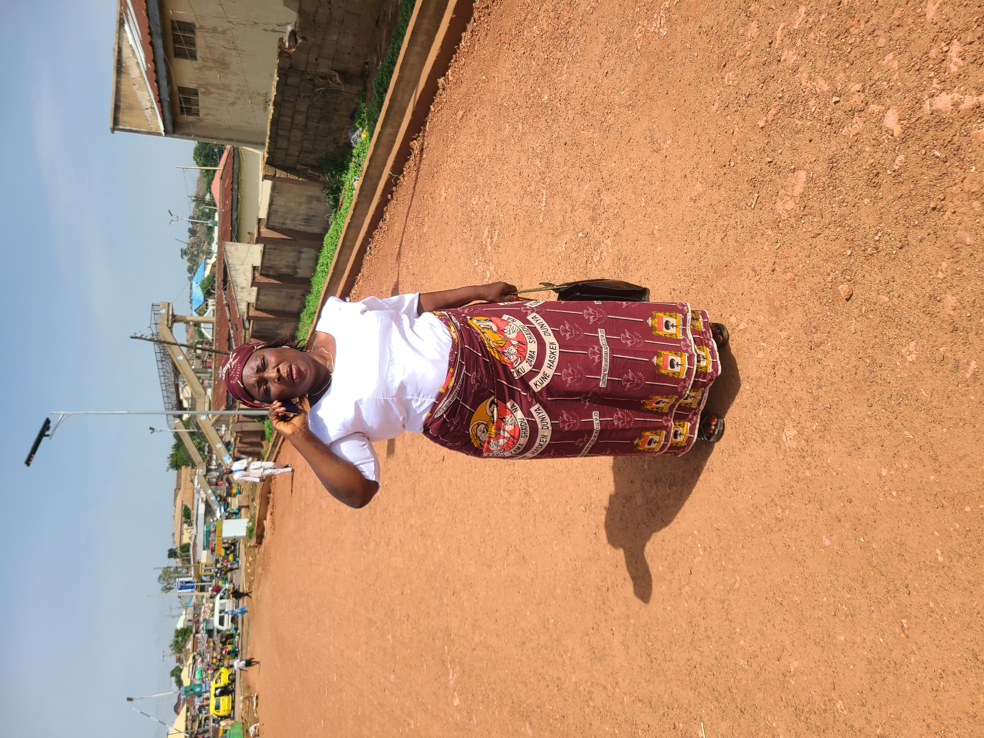 The image captures a street scene in Jos, Nigeria, on a bright, sunny day. In the foreground, a woman is standing and speaking on a cell phone. She is wearing a white, short-sleeved top and a patterned maroon wrap skirt. She also has a maroon headscarf and is carrying a dark-colored bag. The ground is a reddish-brown, dusty dirt road, and a distinct shadow is cast by the woman.
In the background, a busy street is visible with various vehicles, including yellow minibuses and a white van. Pedestrians are also present, and some are walking. Several buildings line the street, some with corrugated metal roofs and others with more traditional construction. A pedestrian bridge is visible in the middle ground, and streetlights are also present. The overall environment appears to be an urban or semi-urban setting with some vegetation along the sides of the road. The sky is clear and blue.
