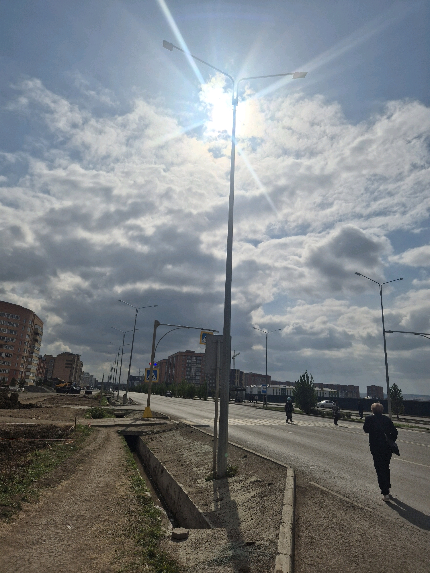 The image captures an outdoor urban scene under a bright, partly cloudy sky, with the sun prominently visible and causing lens flare. The perspective is from a dirt path alongside a road, looking towards a street with multiple lanes of asphalt. The path has a drainage ditch running parallel to it.
On the left side of the road, there are construction activities. Piles of dirt and debris are visible, along with some orange construction fencing. A yellow excavator is parked in the background. Further down the road, multi-story residential buildings with reddish-brown facades line the street.
The road itself is busy with traffic, though not congested. Several cars are visible, and a few pedestrians are crossing the street or walking on the sidewalk. One woman in a dark jacket and pants is walking away from the camera on the right side of the road. Another pedestrian, a child wearing a blue helmet, is crossing the street in the distance. Traffic lights and pedestrian crossing signs are present.
Streetlights are mounted on tall poles along the road. The sky is a mix of blue sky and fluffy white and dark grey clouds, suggesting dynamic weather conditions. The overall impression is of an active urban environment undergoing development, with everyday life continuing amidst construction. The location is identified as Kokshetau, Kazakhstan.