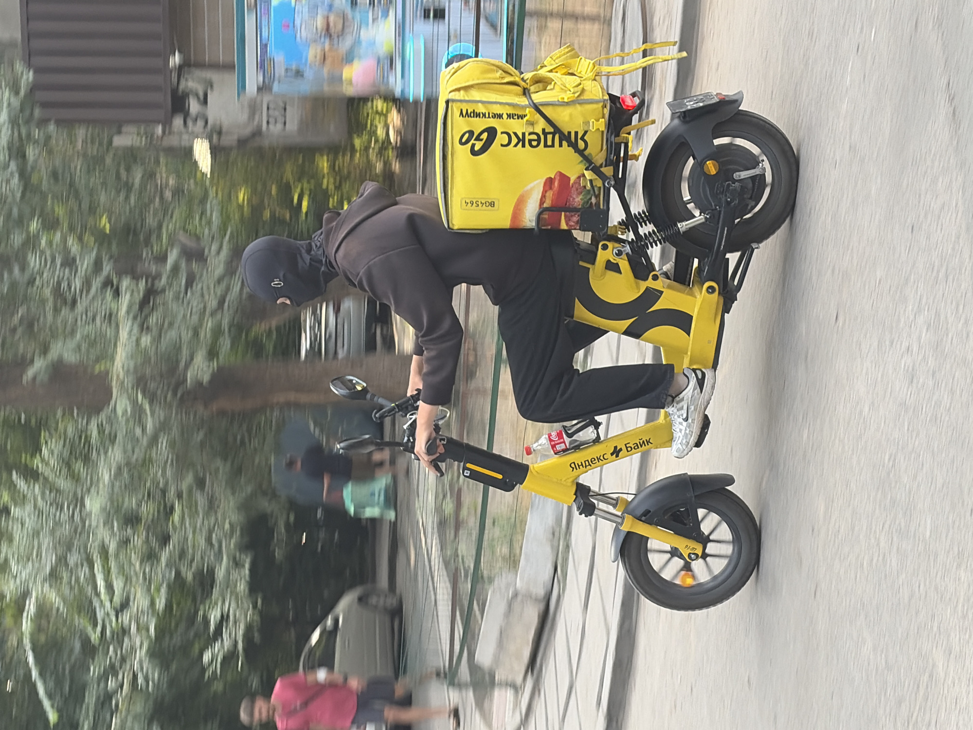 A person wearing a black balaclava and a brown hoodie is riding a yellow electric scooter on a paved road. The scooter has a large yellow insulated bag on the back, branded with "Yandex Go" and an image of a burger. The rider is wearing black pants and white sneakers. A bottle of Coca-Cola is attached to the scooter's frame. In the background, trees and buildings are visible. There are other people and vehicles in the background, but they are out of focus. The scene appears to be in a city.
