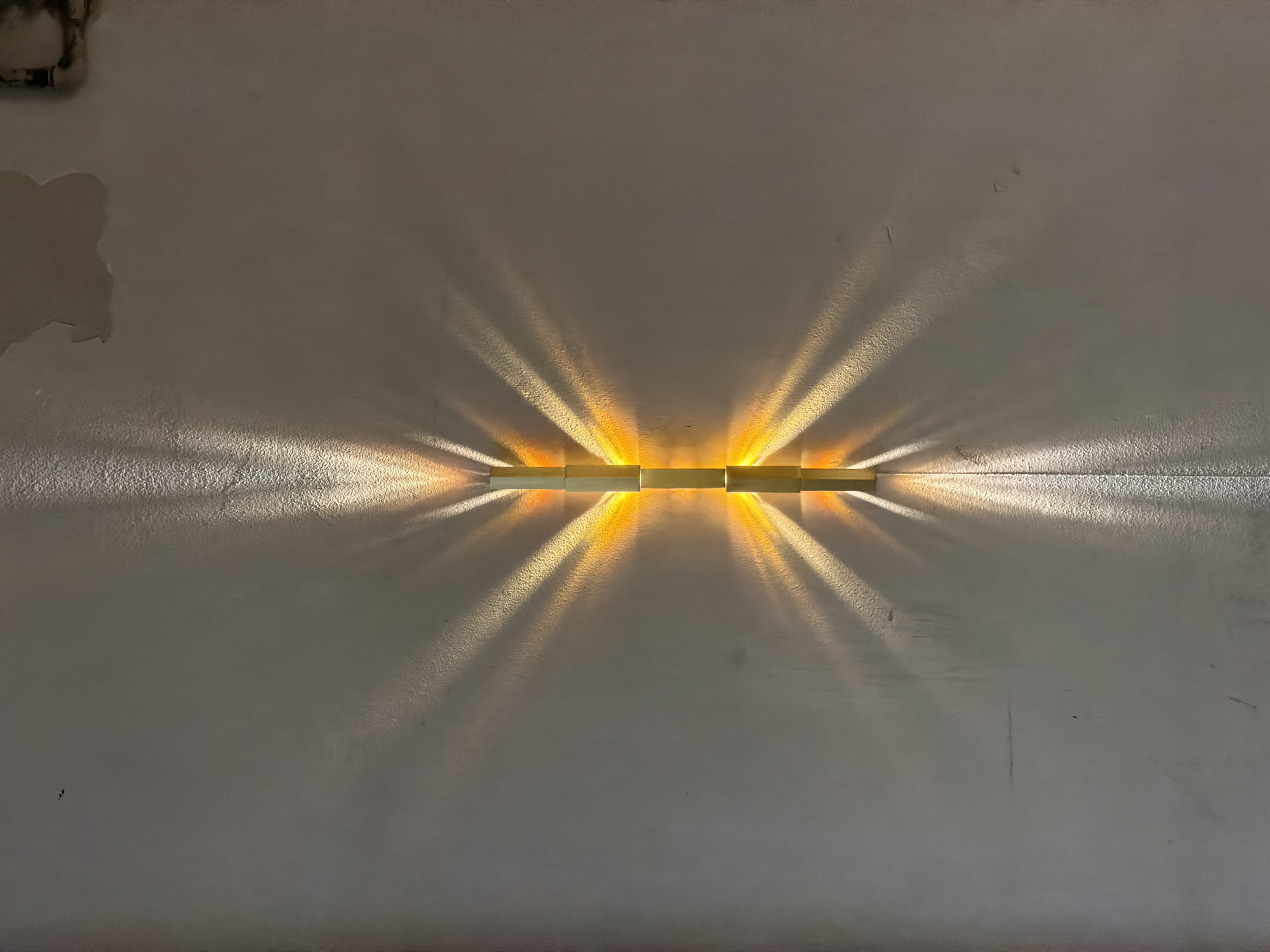 A horizontal light fixture, composed of rectangular segments, is mounted on a textured, off-white wall. The fixture emits bright, focused beams of light upward and downward, creating a radial pattern of illumination on the wall. The wall shows signs of wear, including a patch of missing paint in the upper left corner and various subtle marks and imperfections across its surface. The light fixture and wall are situated in what appears to be an indoor space. There is no indication that the photo was taken in Milan, Italy.
