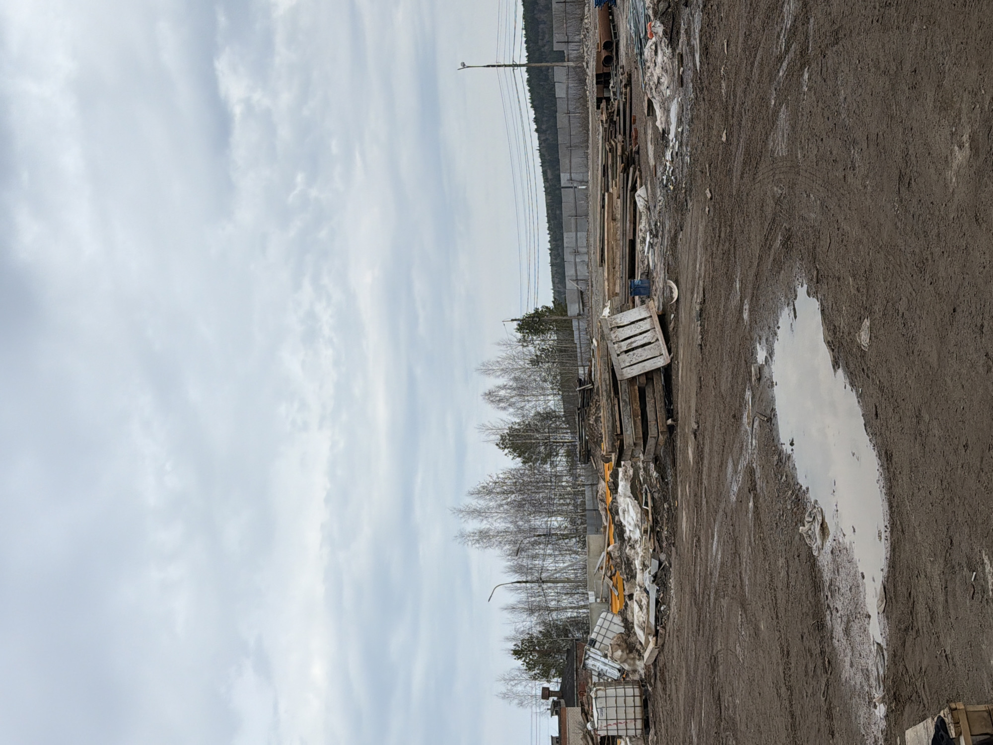 The image depicts a muddy, industrial-looking area in Chaikovsky, Russia. To the left of the dirt road is a collection of discarded items, including wood pallets, plastic containers, and construction debris. Further back, the ground has some snow cover. Trees are visible in the background, with a concrete wall and electrical power lines behind them. The sky is overcast. A large puddle fills a pothole in the foreground.