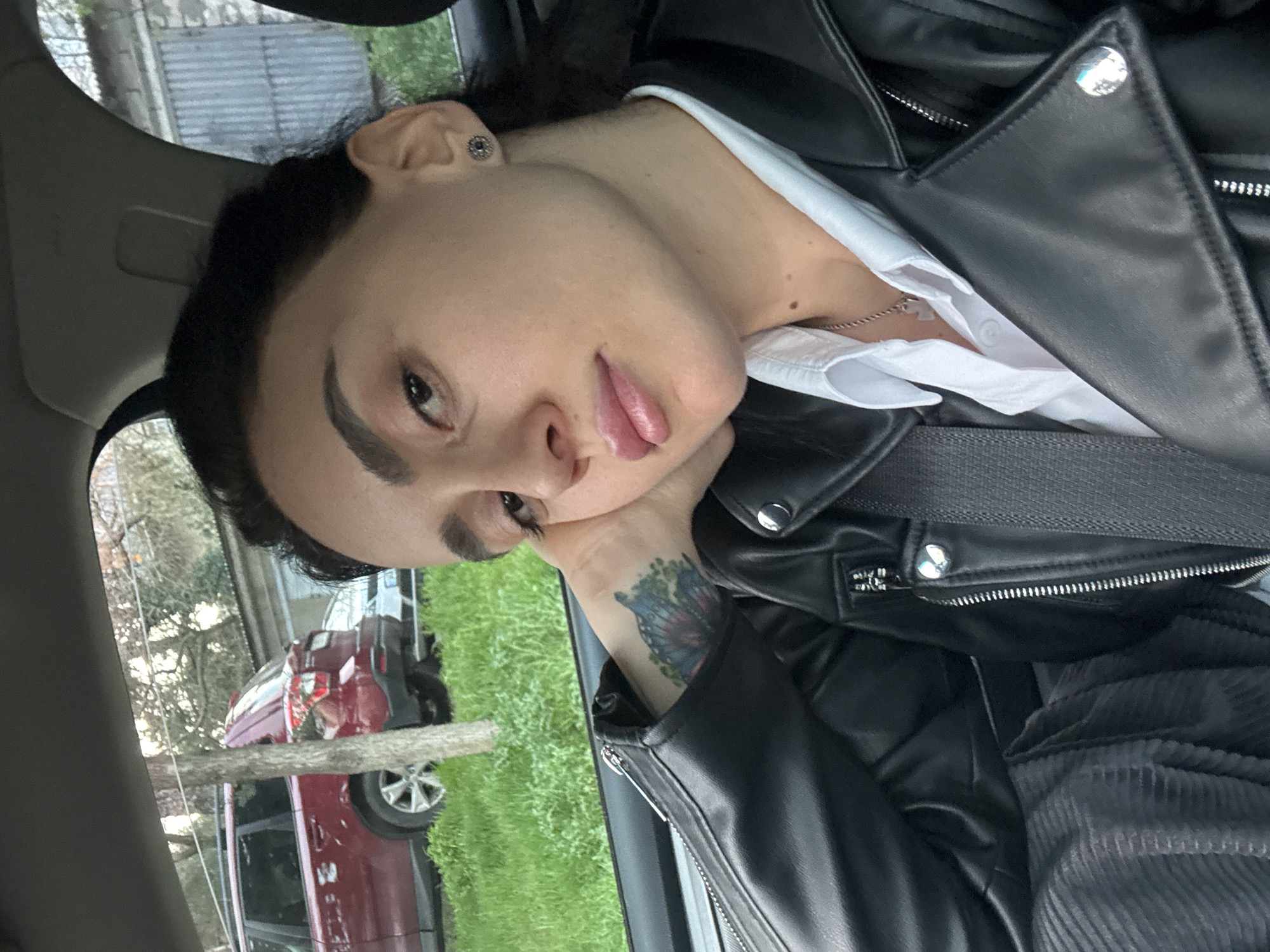A person is posing for a selfie inside a car. The person has fair skin, dark hair, and is wearing a black leather jacket over a white collared shirt, with a seatbelt across their chest. Their head is resting on their left hand, which has a tattoo visible on the wrist. Outside the car window, a lawn and a red SUV can be seen. The scene is likely in Tbilisi, Georgia, based on the general architectural style of buildings and the make of cars visible.