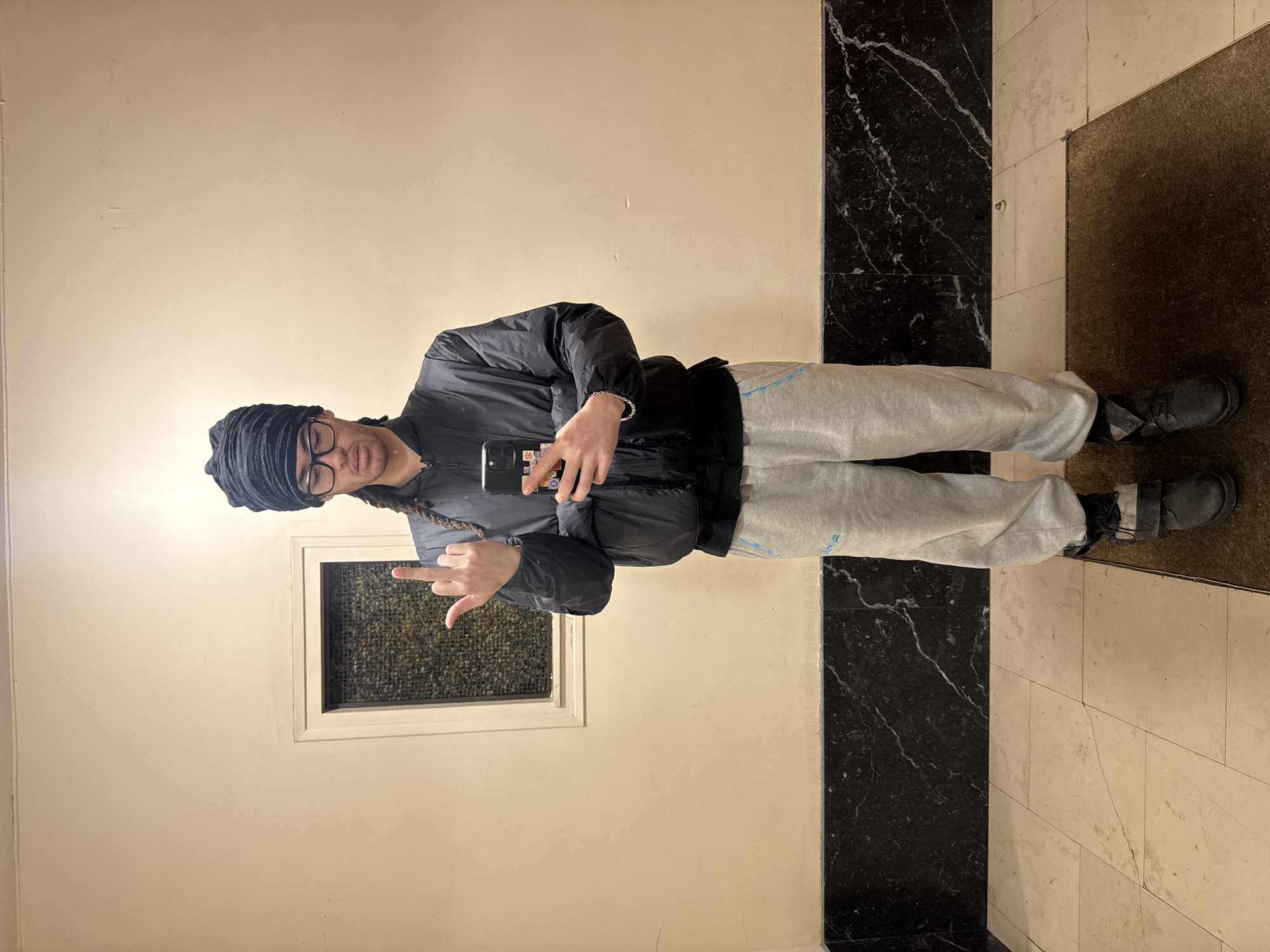 A person wearing glasses, a black head wrap, a black jacket, and grey sweatpants is taking a selfie in front of a wall that has a small window. The person is also wearing black shoes. The floor is tiled in a checkered pattern, with a black marble strip along one side. A brown mat is also visible on the floor. The location appears to be indoors. There is no information to confirm the location is Paris, France. The person is extending the middle finger to the phone, and is holding the phone in their left hand.