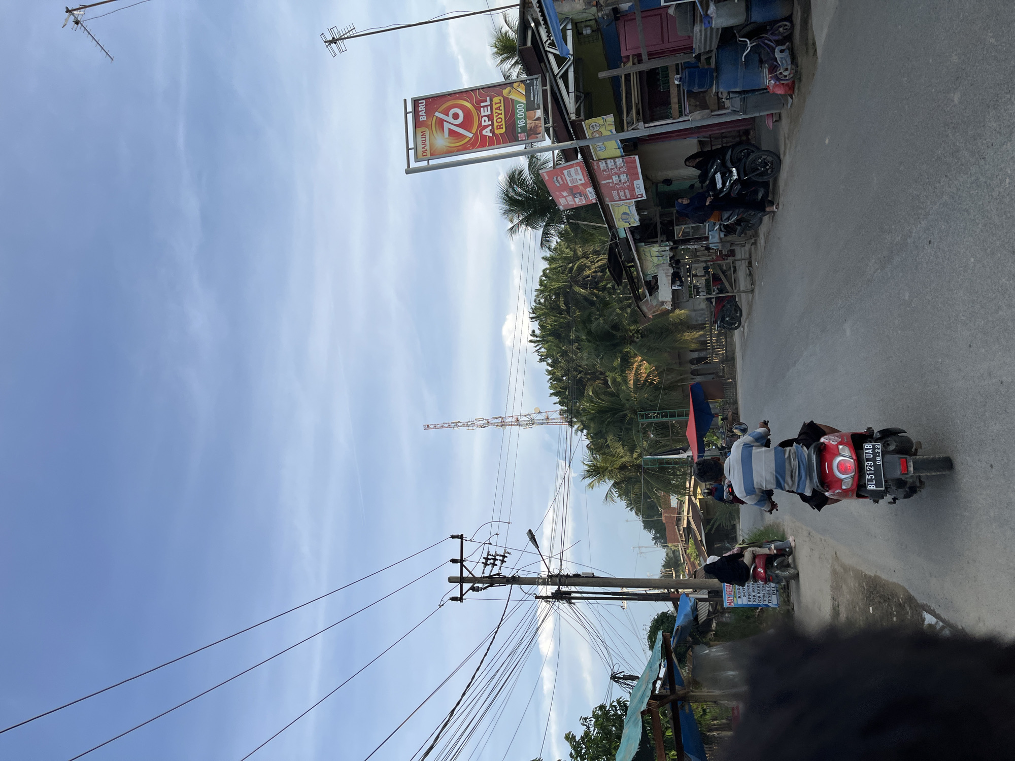 In Alurcucur, Indonesia, a person rides a red scooter down a paved road. The scooter is a red Honda Beat with a license plate reading BL 5129 UAB, dated 08-22. The rider wears a striped blue and white shirt and black shorts. To the right of the road, there is a building with a tall, red and white banner advertising "DIARUM BARU 76 APEL ROYAL". Several other signs and structures line the road, including a utility pole with numerous wires and a distant cell tower. Tropical trees, including palm trees, are visible in the background. Other people and scooters are present on the side of the road and further down the street. The sky is partly cloudy with a bright blue hue.
