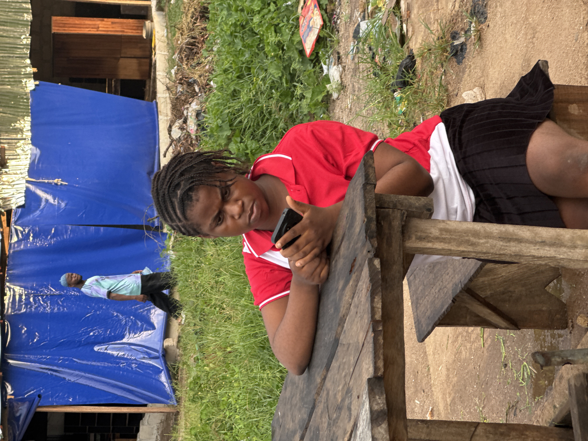 In Ihiagwa, Nigeria, a young woman with braided hair is seated at a rustic wooden table, engrossed in her mobile phone. She is wearing a red collared shirt with white trim and a black pleated skirt. The table is made of rough-hewn wooden planks, and a matching wooden bench is positioned beneath it. In the background, a man in a light blue and teal t-shirt and a light blue cap walks past a large blue tarp that covers a structure. The environment is outdoors, with tall green grass and scattered debris indicating a somewhat unkempt area. A corrugated metal roof is visible above the tarp. The ground is a mix of dirt and patchy grass.