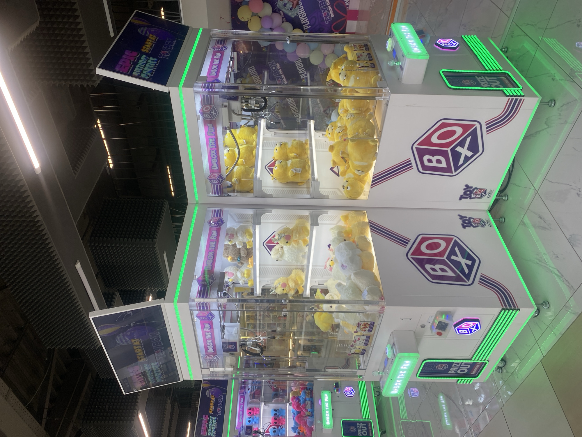 The image shows two brightly lit claw machine arcade cabinets, branded with "BOX" and "TOY BOX" logos, filled with plush toys. The cabinets are white with purple and red accents and feature green LED lighting along the base and on control panels. One cabinet on the left displays various colored alien plush toys and a smaller selection of pink and blue plush toys. The cabinet on the right is filled with yellow and white plush duck toys and yellow stuffed animals. Both machines have large screens at the top displaying colorful graphics and text. The cabinets are positioned side-by-side in what appears to be a shopping mall or entertainment center, with a tiled floor reflecting the green lights. In the background, a wall is decorated with colorful balloons and a sign that reads "PLAYGROUND". The overall environment suggests a commercial entertainment venue.