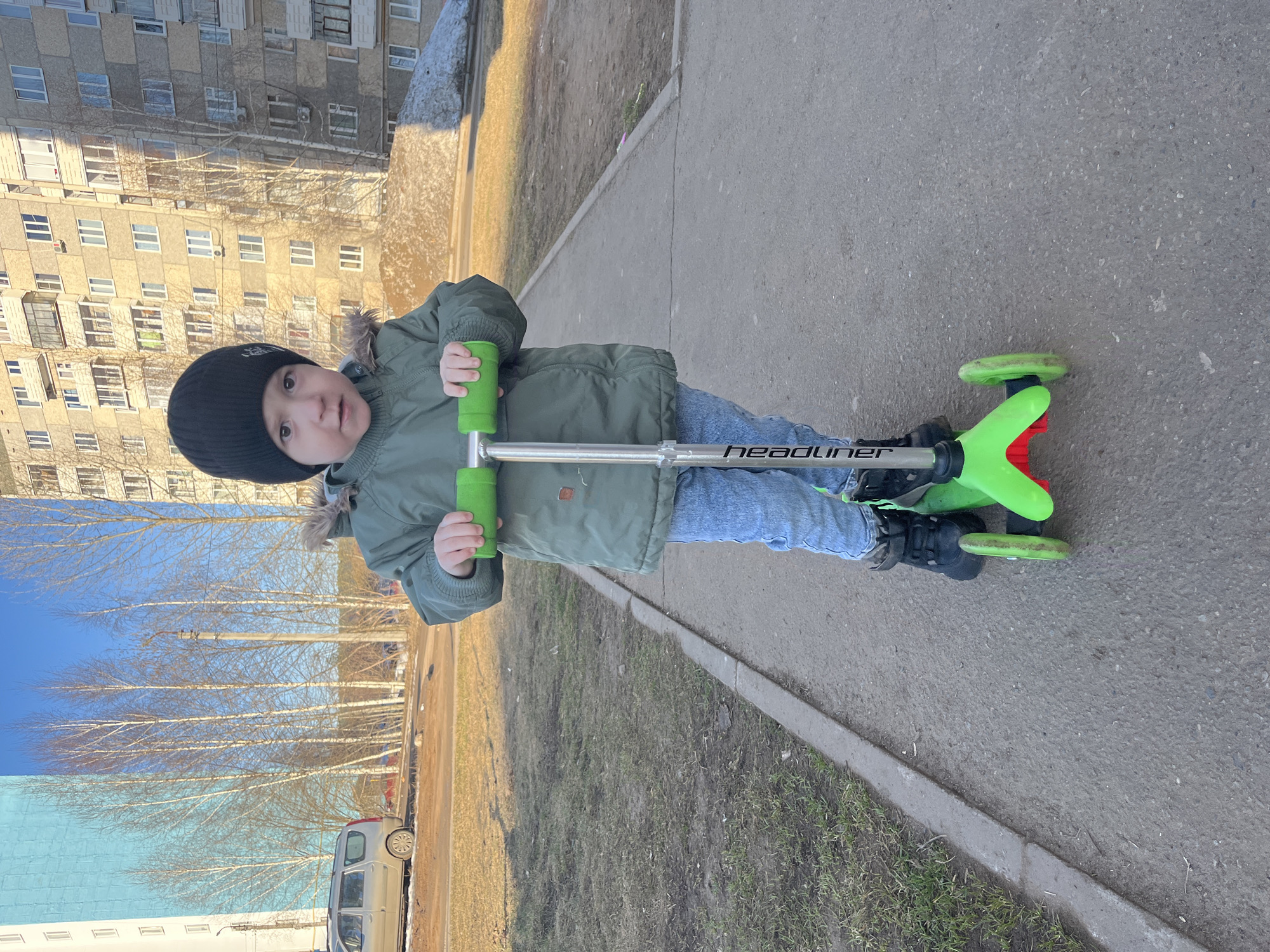 A child, wearing a black beanie, a green jacket with fur trim, and blue jeans, is standing on a green and red scooter on a paved area. The scooter's handlebar reads "HEADLINER." The child is holding the green handlebars and looking at the camera. Behind the child is a grassy area and a building with many windows. In the background, there are bare trees. The location is the city of Chaikovsky, Russia.