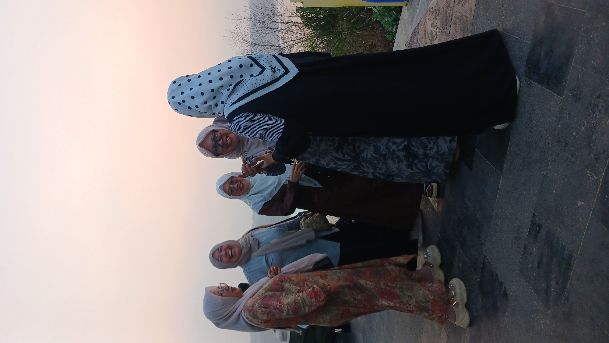 Five women wearing hijabs are gathered on an outdoor paved area, likely a boardwalk or promenade, with a body of water visible in the background. The sky is a pale orange, suggesting it is either sunrise or sunset. The women appear to be interacting and smiling, with one woman in the center laughing heartily. The woman on the far right is wearing a black abaya with a white and black polka dot hijab, and her right hand is raised, possibly holding a phone or taking a picture. Another woman in a floral-patterned dress is on the far left. The ground is paved with dark gray square tiles. The overall impression is of a casual social gathering outdoors. The specific location in Tanjungsari, Indonesia, is not visually discernible from the image itself, but the presence of a body of water and lush vegetation on the right bank indicates a natural or semi-natural setting near the coast or a large river.