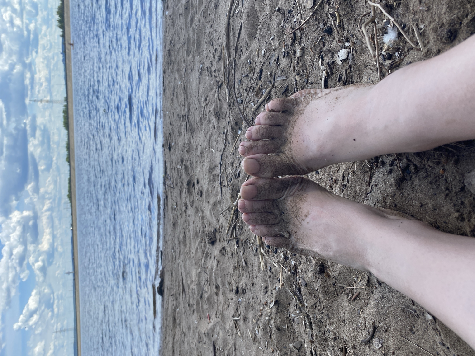 This media file shows a close-up view of a person's feet resting on a sandy shore. The feet are bare and partially covered in sand, suggesting they have been in or near the water. The scene is set by a body of water on the left side of the frame, which appears to be a lake or a large river, reflecting the cloudy sky. The water has a choppy texture, indicating a breeze. The shore is composed of sand and scattered debris, including small sticks and possibly shells.
In the background, a distant treeline is visible, and beyond that, faint structures that resemble power lines or poles can be seen against the sky. The sky is partly cloudy, with fluffy white clouds against a blue background, suggesting it is daytime. The overall mood is relaxed and serene, evoking a sense of being outdoors in nature. The image is oriented such that the feet are in the foreground, and the water and distant landscape recede into the background. The lighting suggests it could be late afternoon or early evening. There is no visible text in the image. The location context is Noviy, Russia.