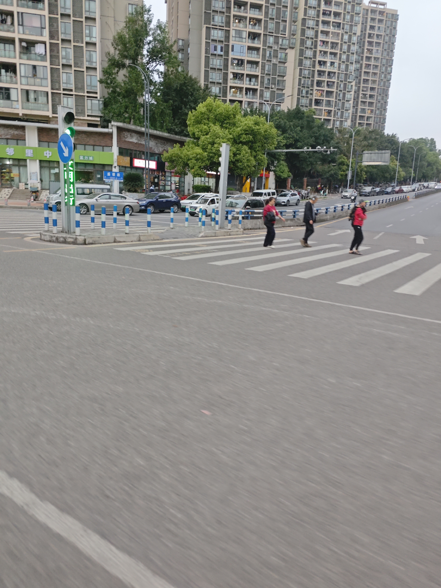 The image depicts a street scene in the Chongqing High-tech Zone, China. Three pedestrians are crossing a zebra-striped crosswalk. Behind them are several cars, a street with more cars parked along the roadside, buildings and trees. The sky is overcast. A traffic light indicates a pedestrian crossing signal. Blue and white bollards border the crosswalk.