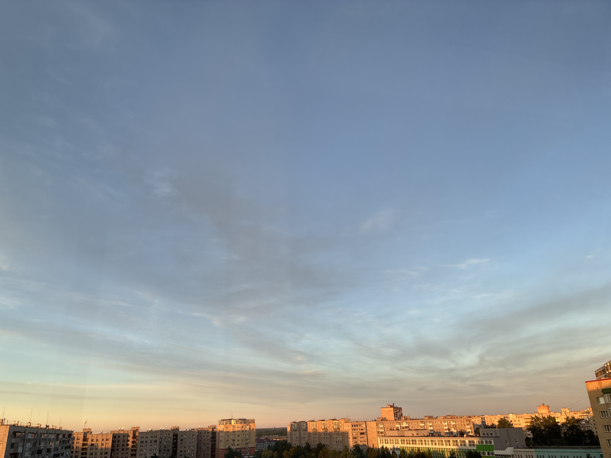 An eye-level shot captures an urban skyline at dusk, showcasing a row of apartment buildings stretching across the frame. The buildings, predominantly gray and beige, vary in height and architectural style, some featuring balconies and windows that reflect the fading sunlight. The sky above transitions from a pale orange hue near the horizon to a soft blue, with wispy clouds adding texture. Trees are visible interspersed between the buildings, their dark foliage contrasting with the illuminated facades. The scene appears to be an aerial view of a residential area within an urban environment.