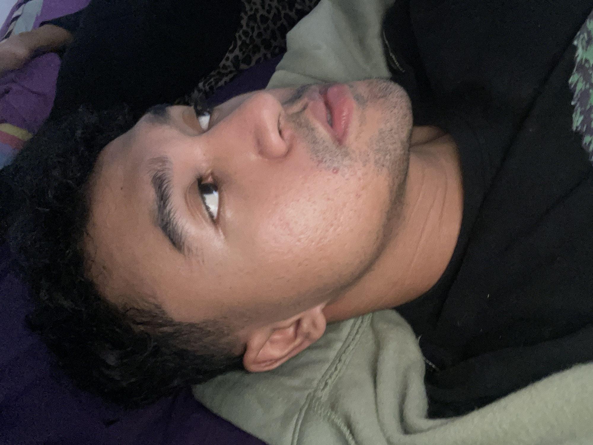 The image features a young adult male lying on his side, looking upwards and towards the right side of the frame. He has dark, curly hair and short stubble on his chin and upper lip. His skin tone is medium brown, and his expression appears neutral and relaxed, with his lips slightly parted.
He is wearing a dark, possibly black, long-sleeved top or hoodie. An olive green fabric, likely a blanket or another part of his clothing, is draped around his head and neck area, showing a ribbed texture. A vibrant purple fabric, potentially a pillow or blanket, is visible behind his head on the left side of the frame. In the upper left corner, a small section of a leopard print fabric is also discernible, suggesting additional bedding or clothing items.
The setting is indoors, likely a bedroom or living space, indicated by the soft furnishings. The lighting is gentle and ambient, creating subtle shadows that highlight his facial contours, consistent with an indoor light source. There are no specific visual cues within the image to indicate the provided context of North Jakarta, Indonesia. No visible text is present in the media file.