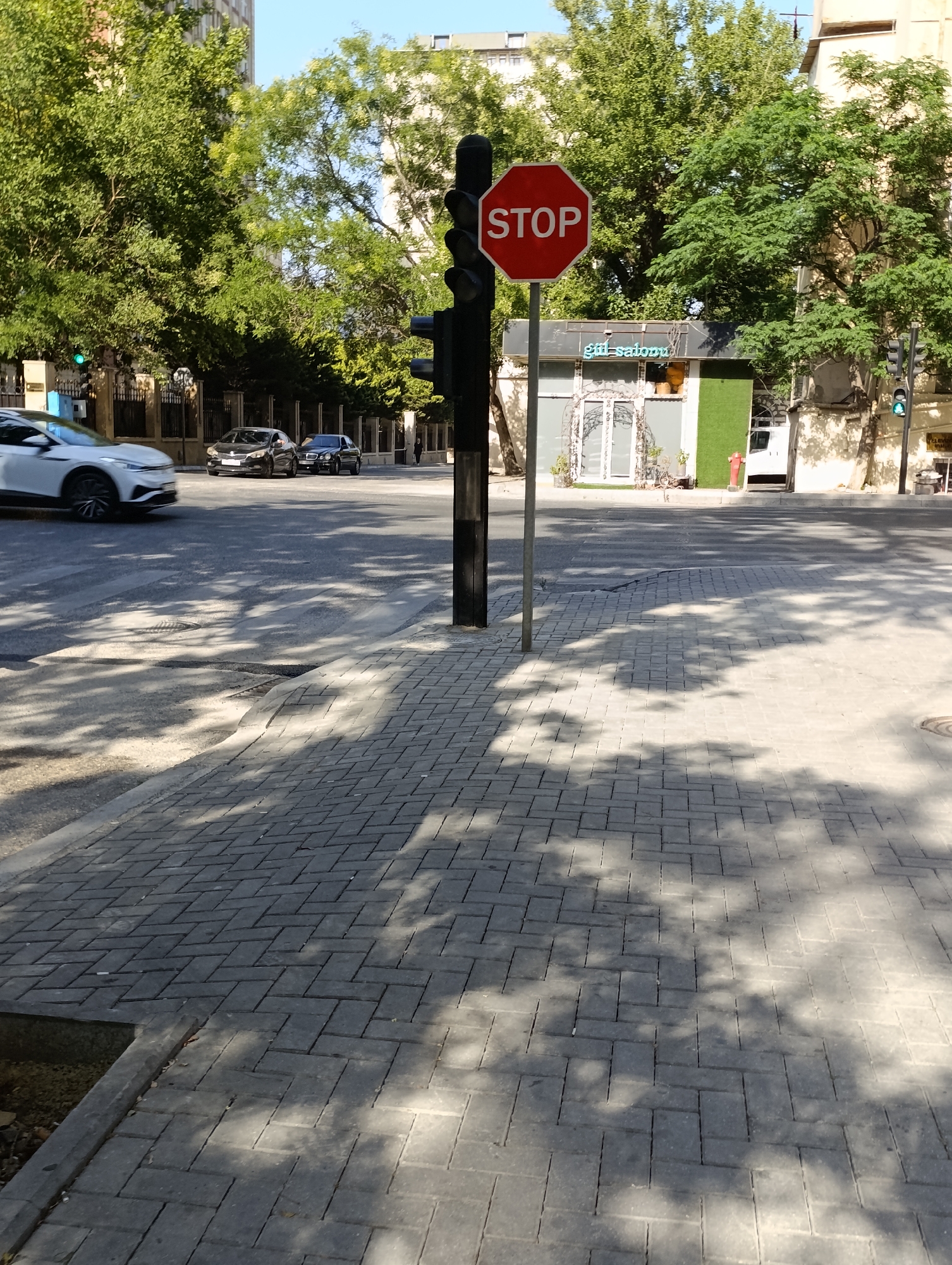 The image depicts a street intersection in Baku, Azerbaijan, under a clear sky on a sunny day. The foreground features a paved sidewalk with a textured pattern of grey interlocking bricks, casting dappled shadows from overhead trees. A concrete curb separates the sidewalk from the road, which is also paved and shows painted lane markings.
Dominating the center of the frame is a traffic light pole with a prominent red octagonal stop sign mounted on it. The stop sign has white lettering that clearly reads "STOP." Below the stop sign, the traffic light itself is visible, with its lower signal light illuminated green.
On the road, several cars are present. A white car is in motion on the left side of the image, appearing to be turning. Behind it, two dark-colored cars are stopped. One is a sedan, and the other appears to be a larger luxury sedan.
In the background, a low-rise building with a modern facade, possibly a shop or cafe, is visible. It has large glass windows and an awning with the text "gül salonu" written on it, suggesting it is a beauty salon. Green trees with dense foliage line the street, and taller buildings are visible in the distance. A red fire hydrant is positioned near the building. The overall scene conveys a typical urban streetscape in Baku during daylight hours.