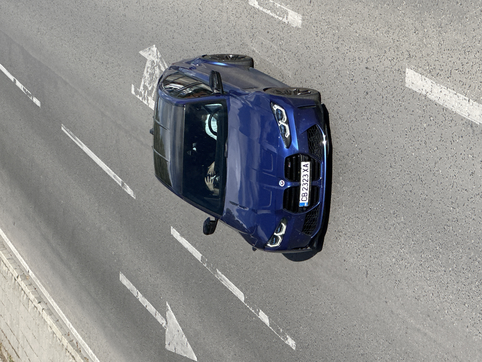 An aerial perspective shot shows a blue BMW sedan driving on a grey asphalt road. The road has white directional arrows painted on it. The license plate on the front of the car reads "CB 2323 XA." The location is likely in Veliko Tarnovo, Bulgaria, based on the license plate format. The road also features a concrete barrier on one side. A person can be seen in the driver's seat.