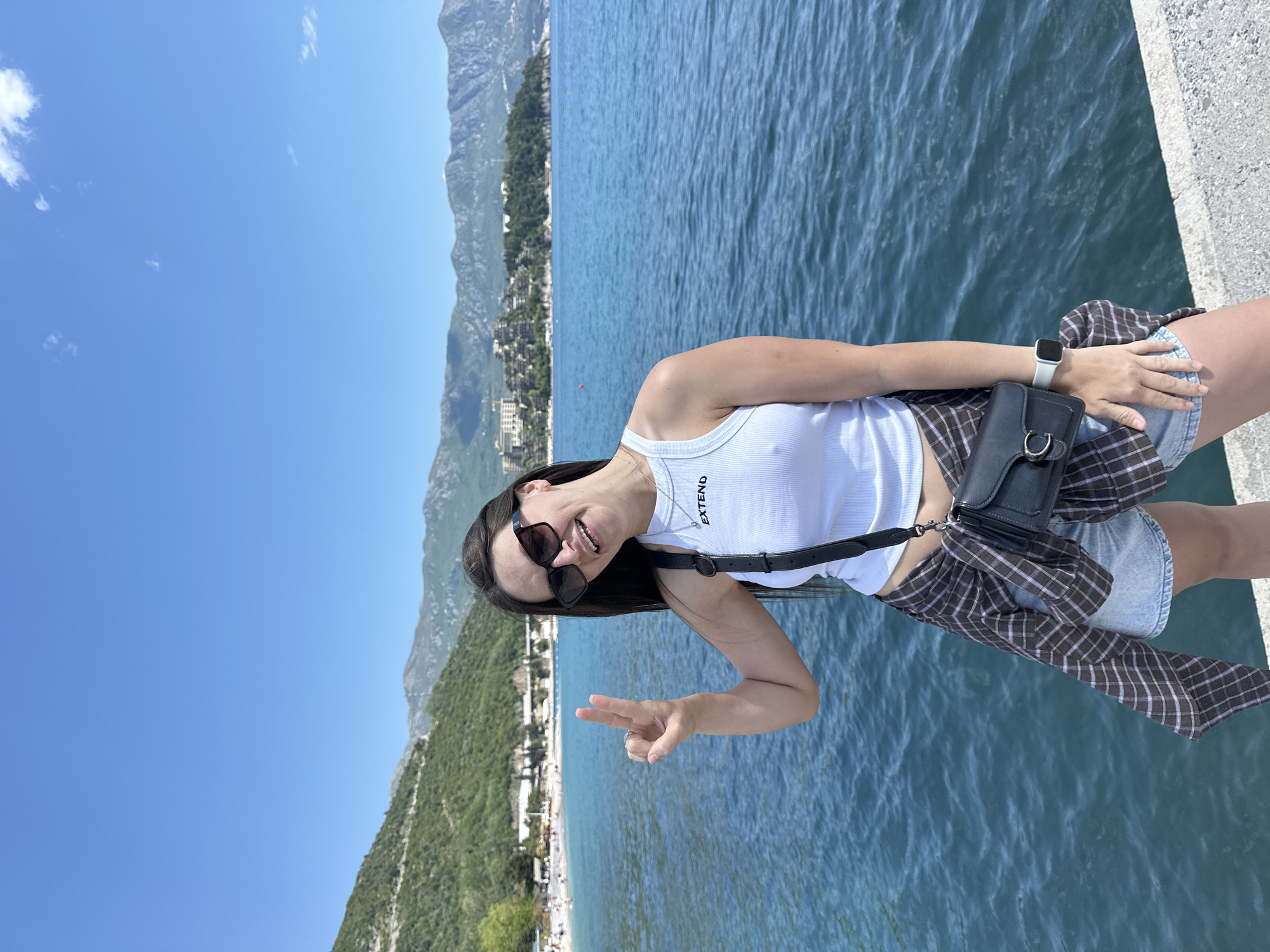 A woman is standing on a concrete edge overlooking a body of water, possibly the Adriatic Sea. She is wearing a white tank top with the word "EXTEND" on it, denim shorts, and a black and brown plaid shirt tied around her waist. She has a black purse with a silver buckle slung over her shoulder. She is wearing sunglasses and a white smartwatch. She is smiling and holding up a peace sign. In the background, mountains and a coastal town can be seen. Based on the surrounding landscape, it is likely the photo was taken in or near Budva, Montenegro.