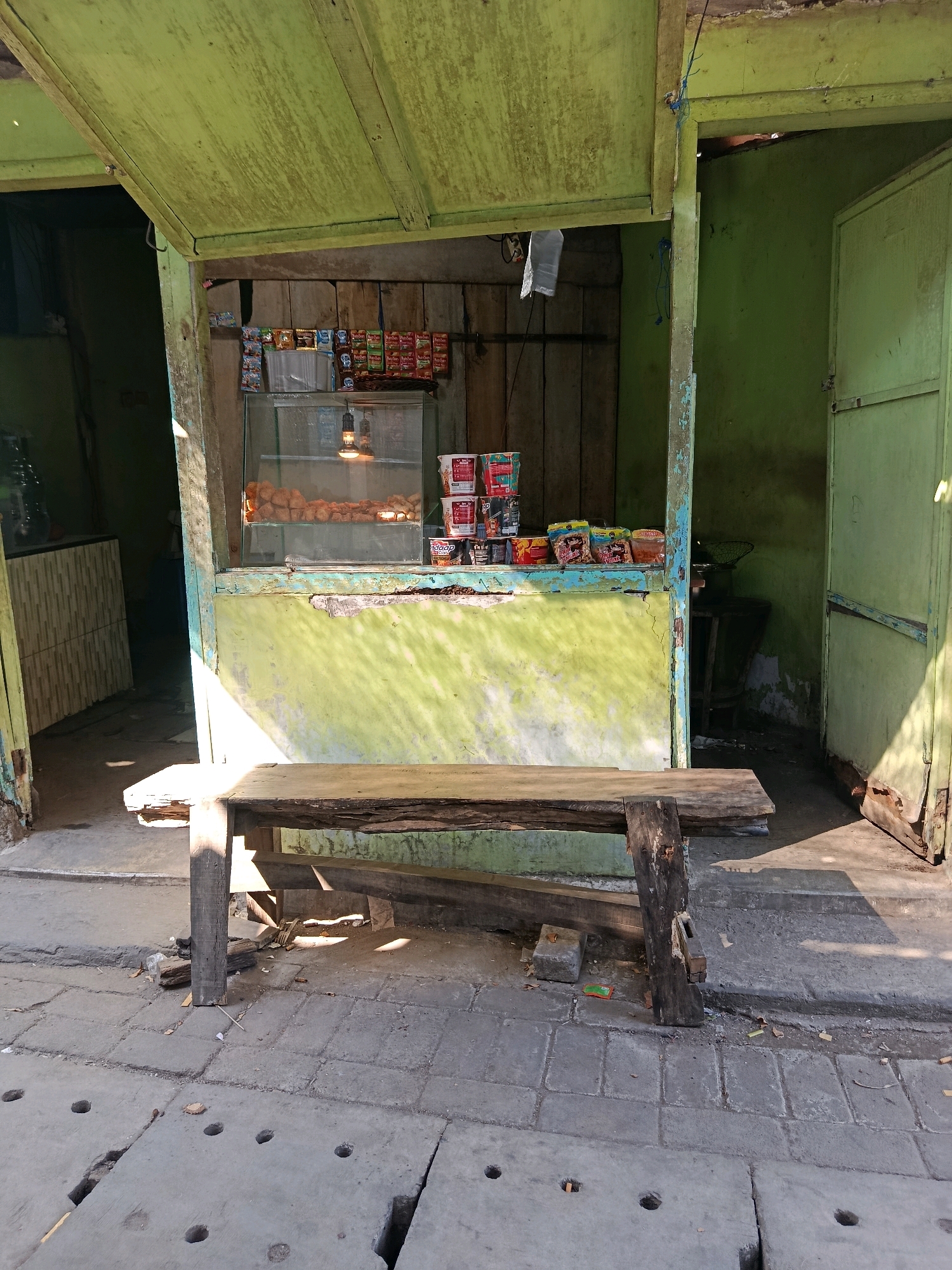 The image shows a small, weathered, open-fronted food stall in Jobokuto, Indonesia. The stall has a lime green painted exterior that is peeling in several places. Inside, shelves are stocked with various packaged snacks and drinks. A glass display case in the center contains what appear to be fried food items, illuminated by a single bare bulb. In front of the stall, a rustic wooden bench sits on a paved area with some cracks and holes. The ground is a mixture of concrete slabs and rough pavement. Sunlight casts strong shadows across the scene, indicating it is daytime. The stall appears to be unattended, and there are no people visible. The overall impression is one of a simple, perhaps aging, local business.