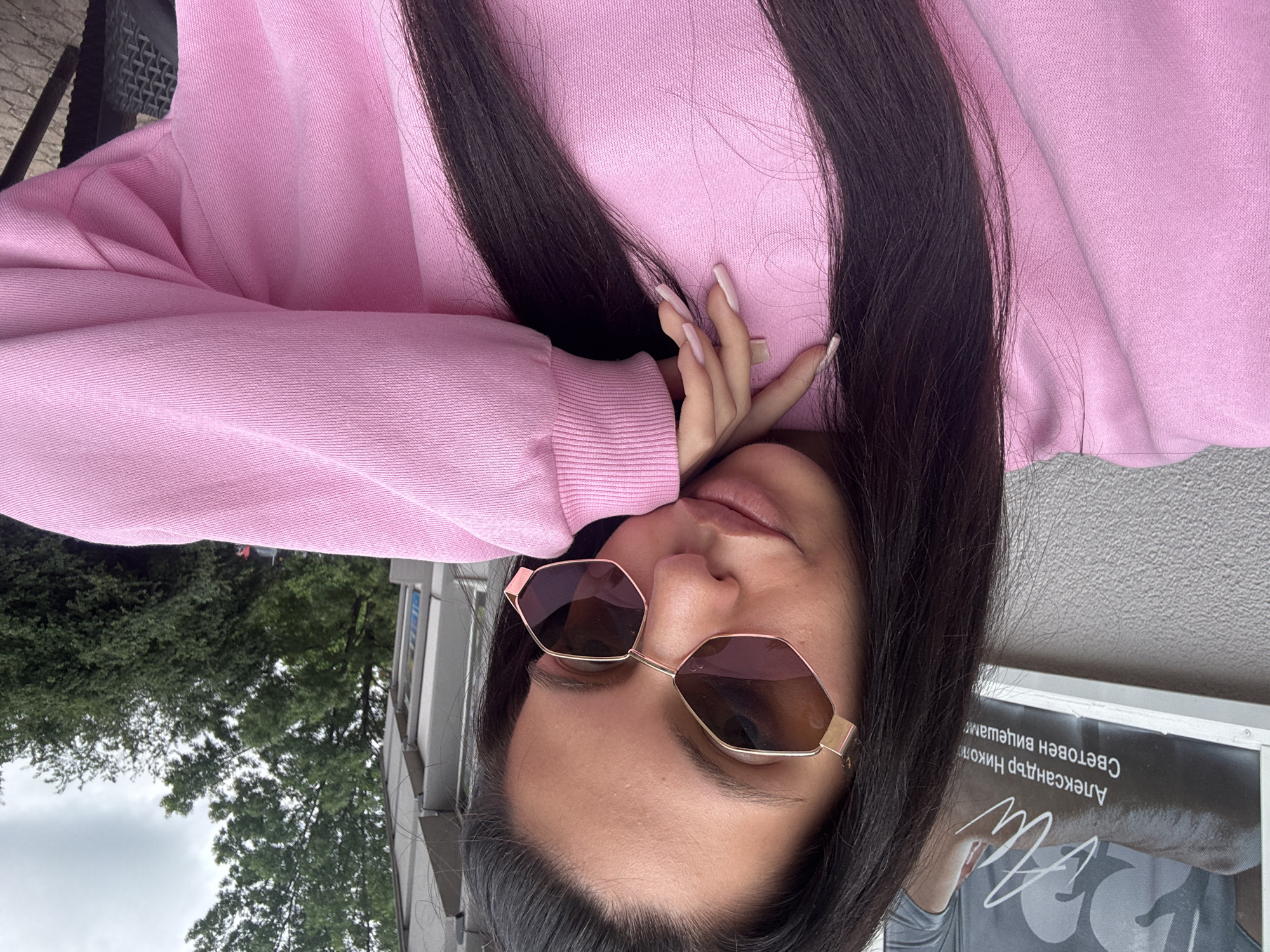 In a selfie taken outdoors in Sofia, Bulgaria, a young woman with long dark hair and light skin is seen wearing a pink sweatshirt and distinctive geometric sunglasses with pink-tinted lenses. Her right hand, with long light pink nails, rests on her chin and cheek. The background shows a textured grey wall with some signage featuring Cyrillic text, a building, and green trees under an overcast sky. The overall impression is casual and posed.