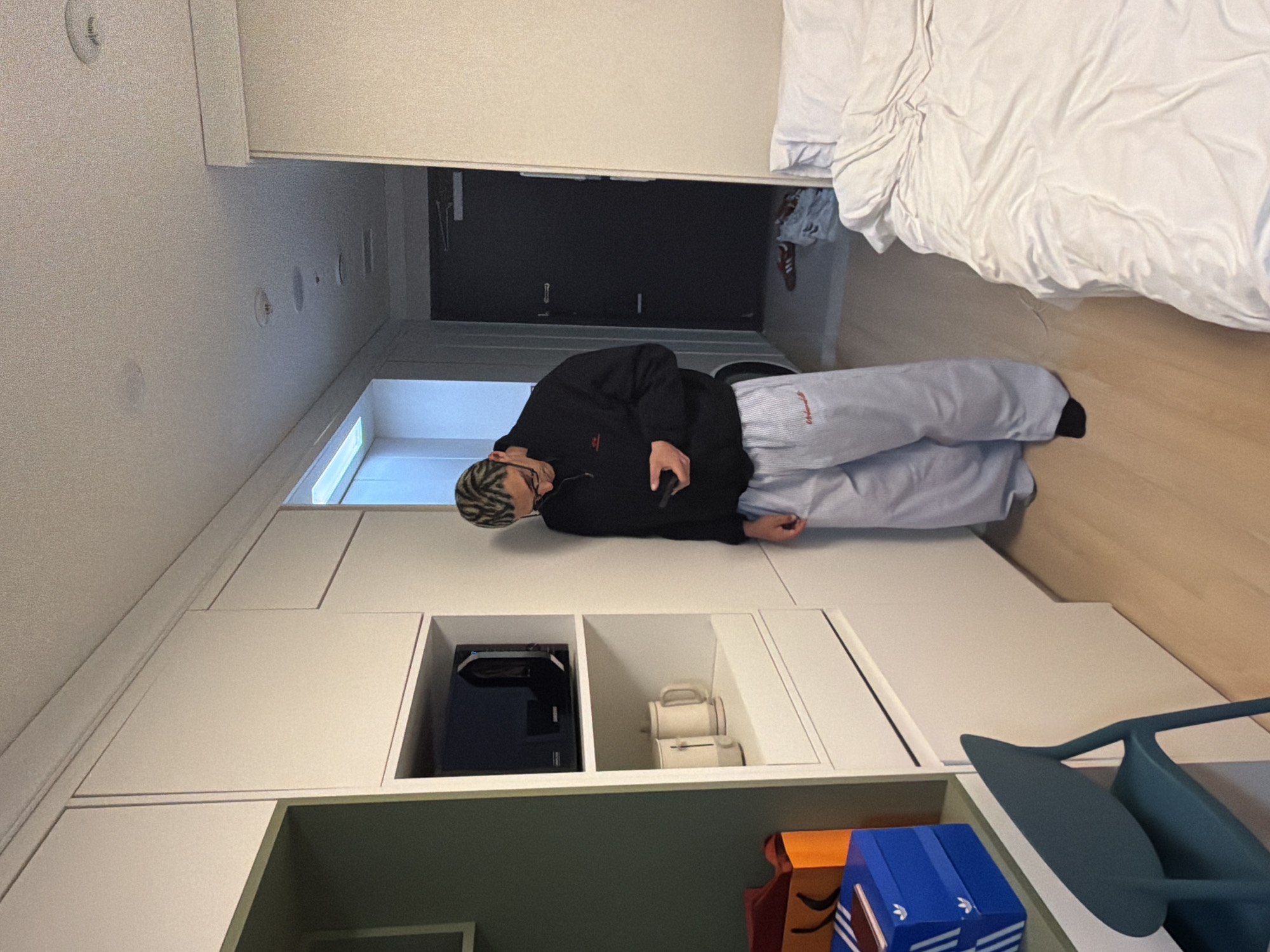 A man is lying on a surface in what appears to be a room in Seoul, South Korea. The room features built-in white cabinets with a microwave and a toaster visible in the shelving. Below these cabinets, there are boxes with blue and orange hues. To the right of the boxes is a blue chair. The man is wearing a black sweatshirt and light-colored sweatpants with dark socks. He is holding a remote control in his hand. The room also includes a bed with white linens, positioned on the right side of the frame. A door, seemingly black, is in the background.